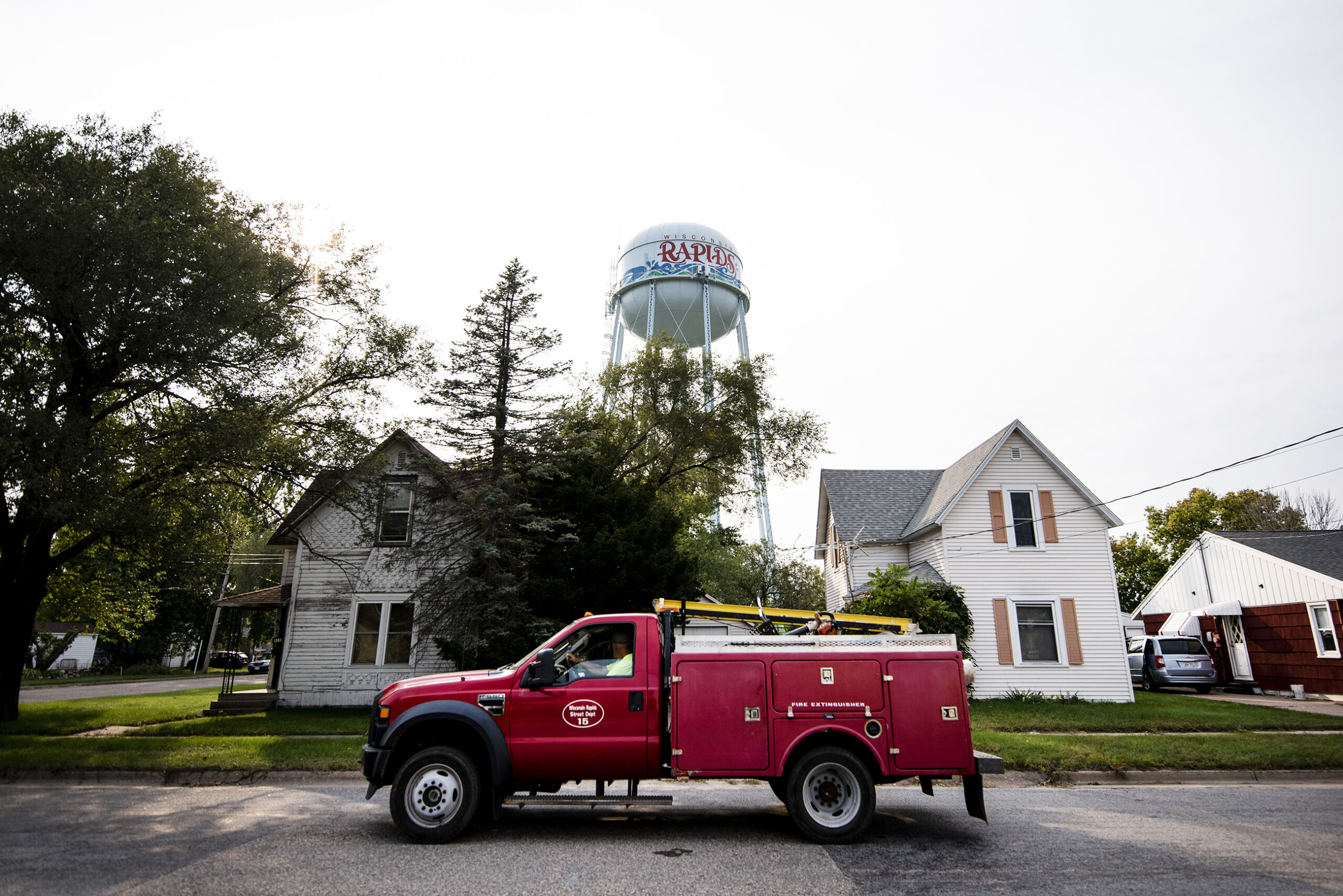 a bright red truck passes by two homes and a water tower that says 