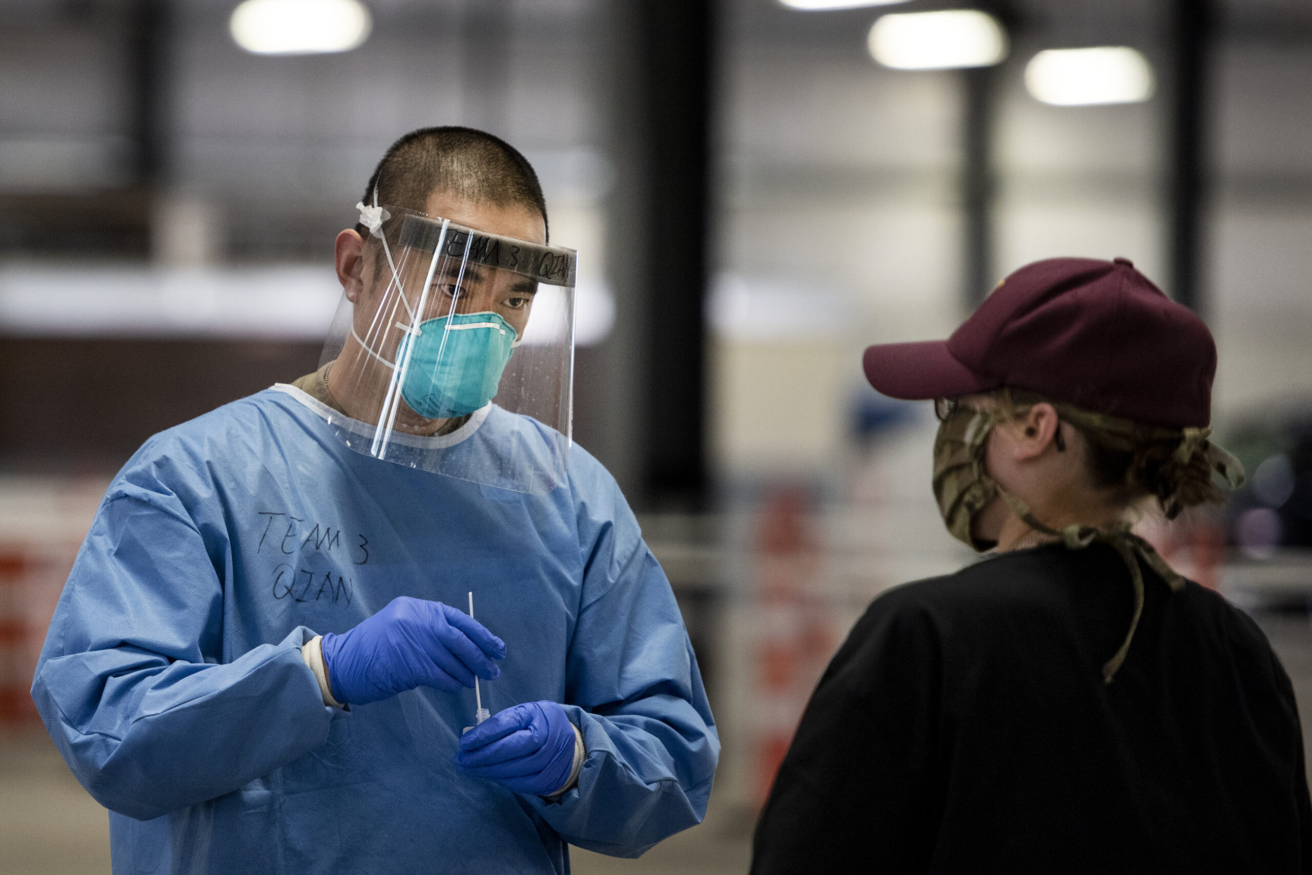 A man in a face shield, mask, blue gown, and gloves inserts a test swab into a container