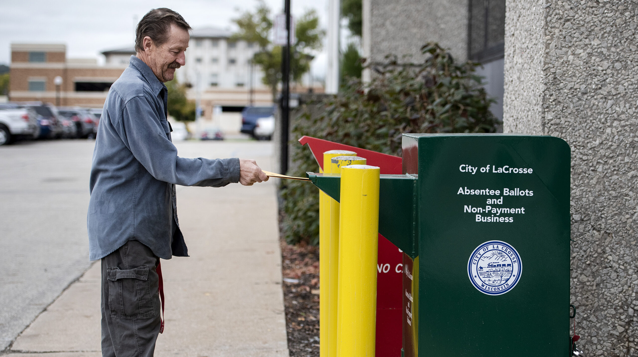A man places a ballot in a yellow envelope into a green drop off box that resembles a public mail box.