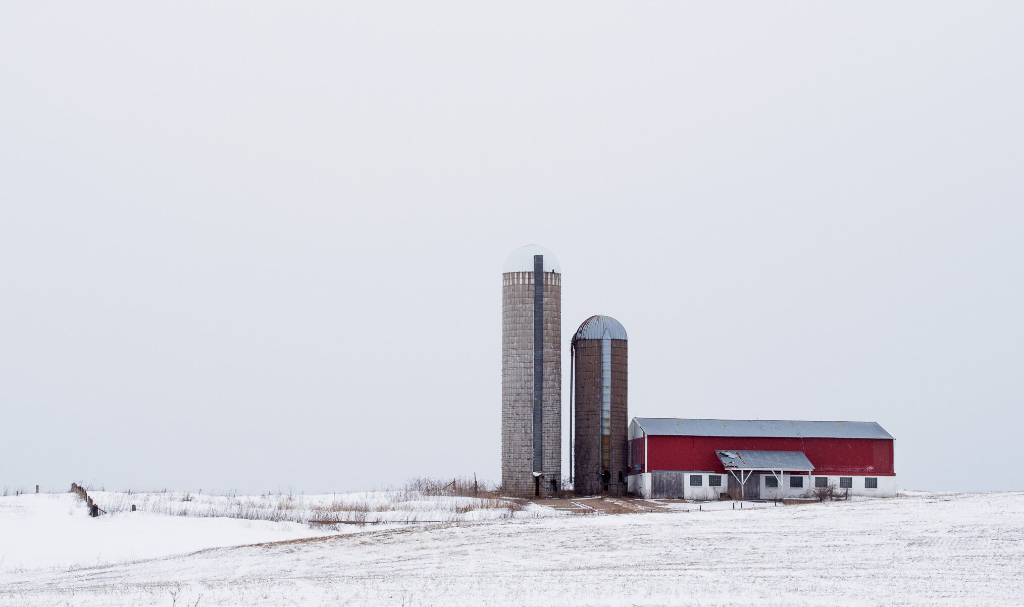 A red barn with two silos stands in a snowy landscape under a gray sky.