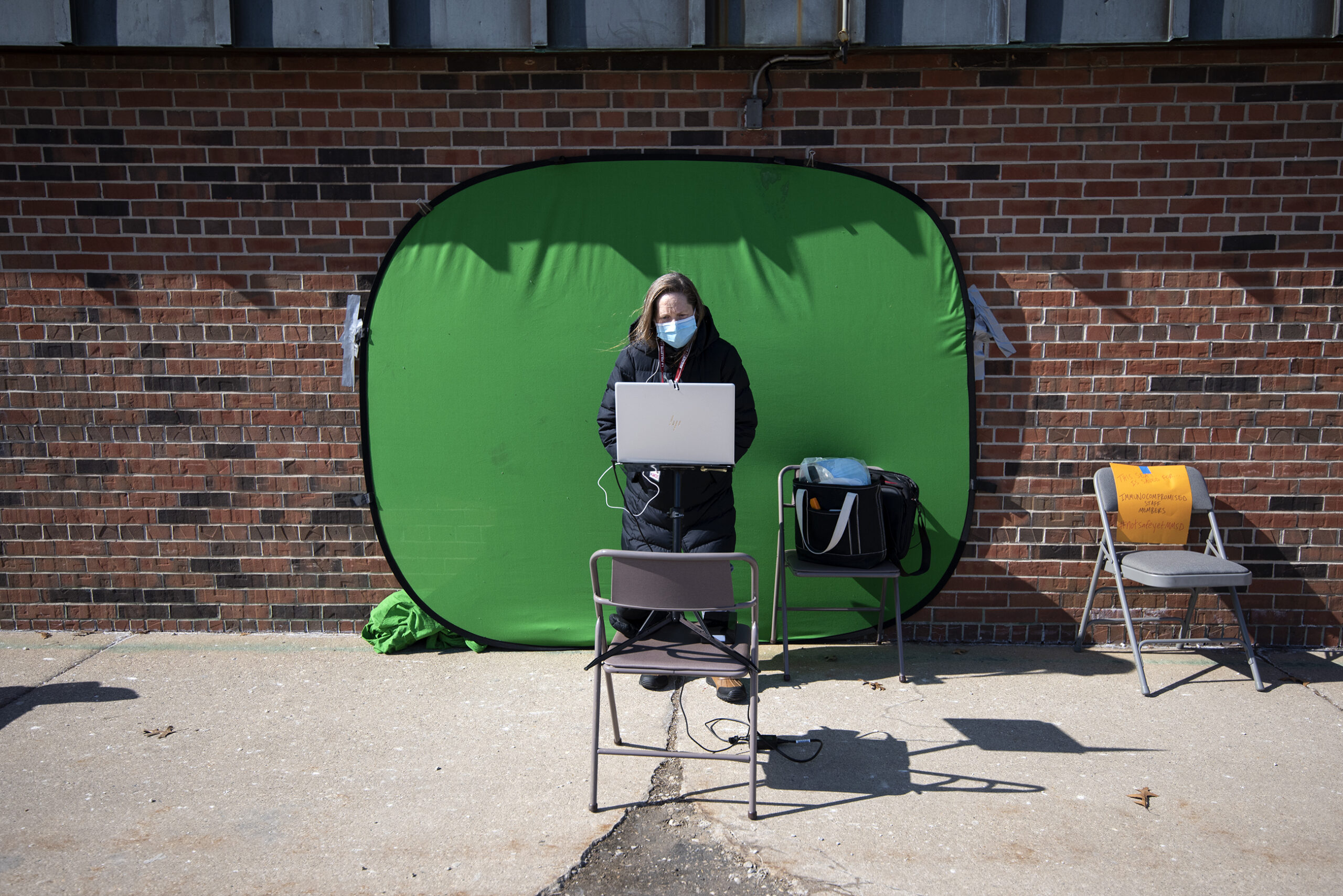 A green screen is seen behind a teacher standing at a laptop.
