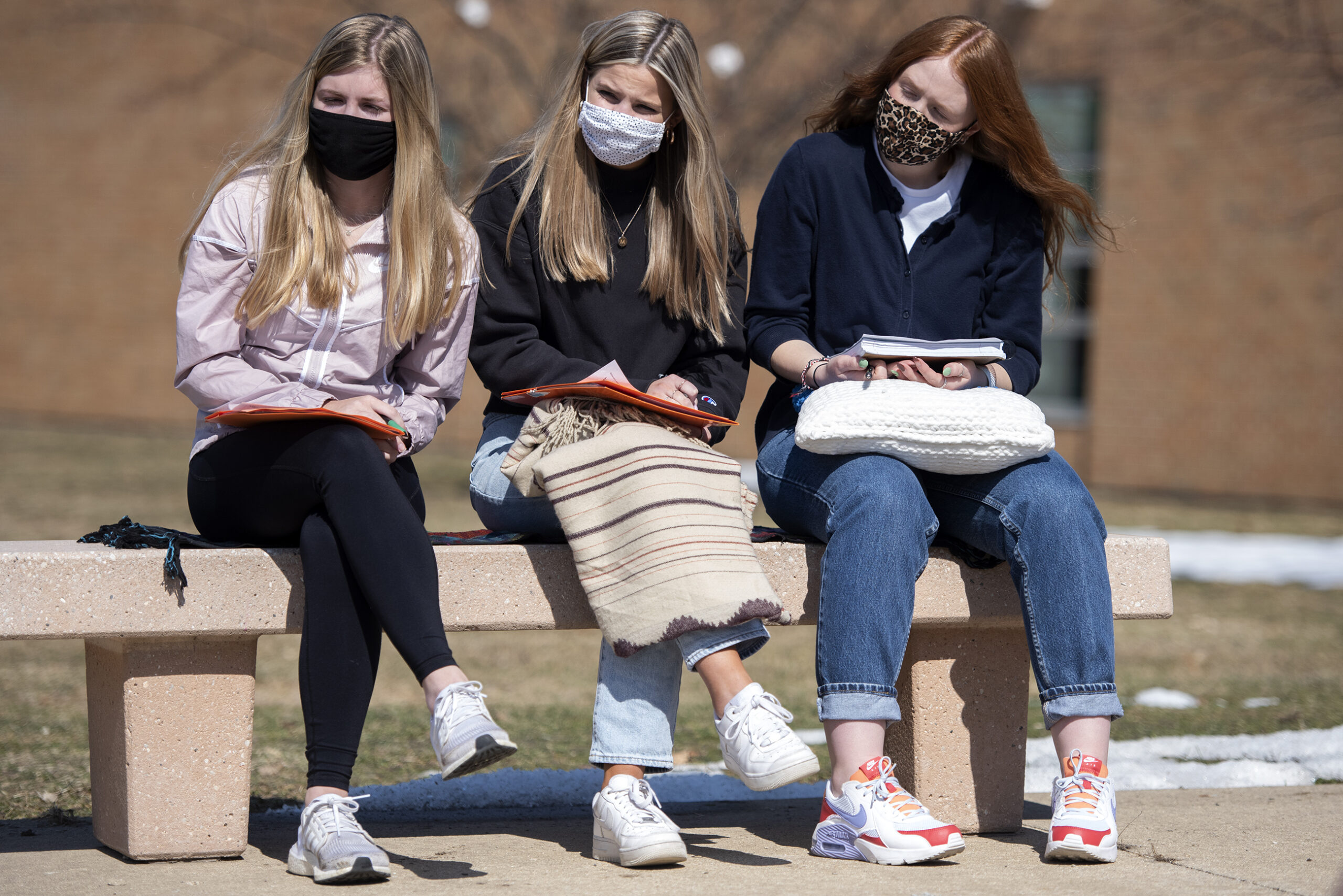 Three girls with long hair sit on a bench outdoors holding notebooks and wearing face masks.
