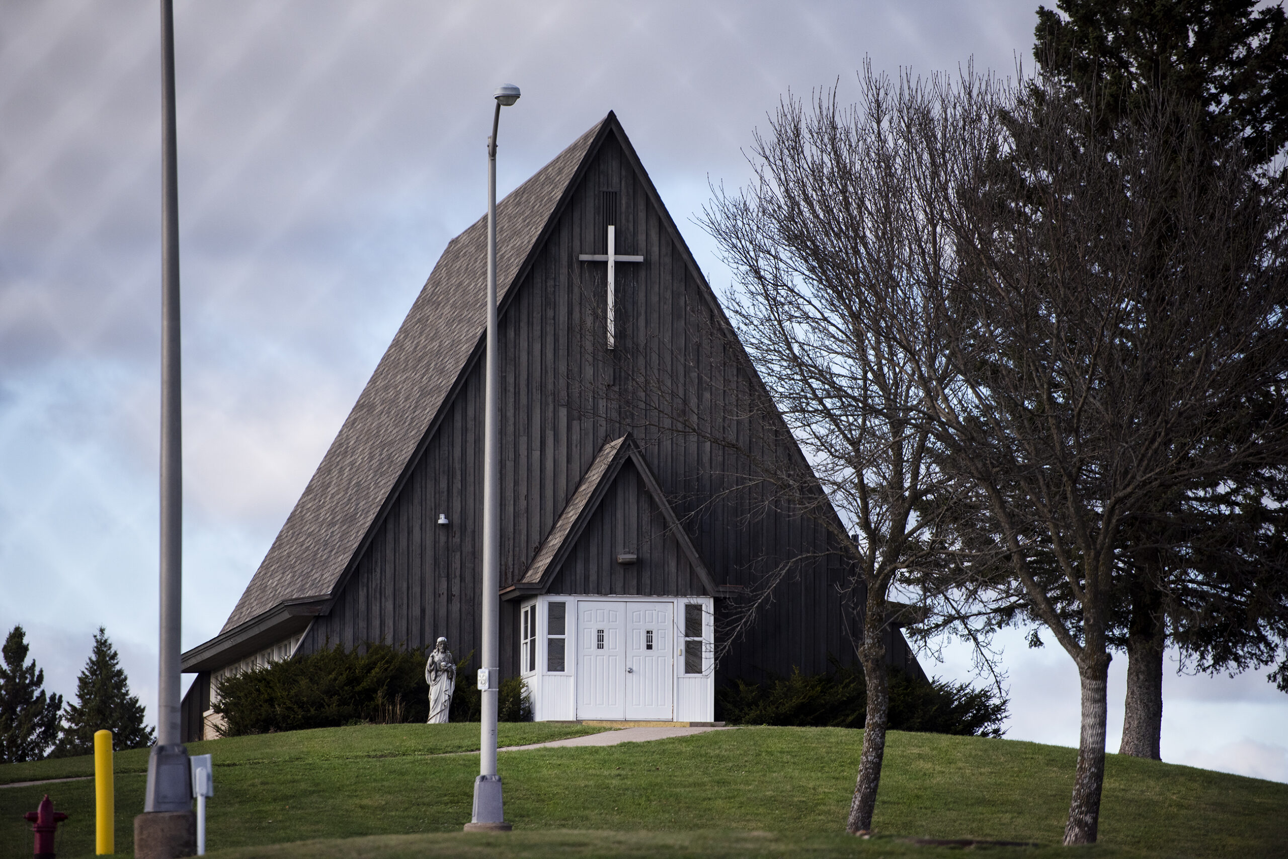 A dark wood chapel has a steep roof. A cross is displayed outside.