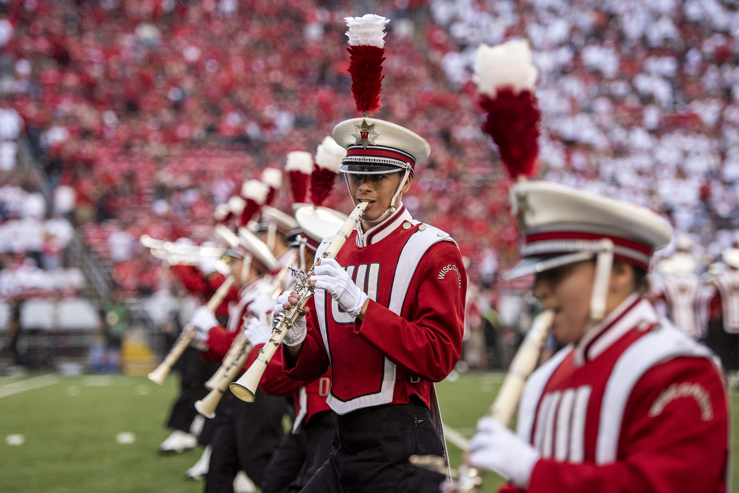 Marching band members form a line across the field while wearing red and white uniforms with plumes.