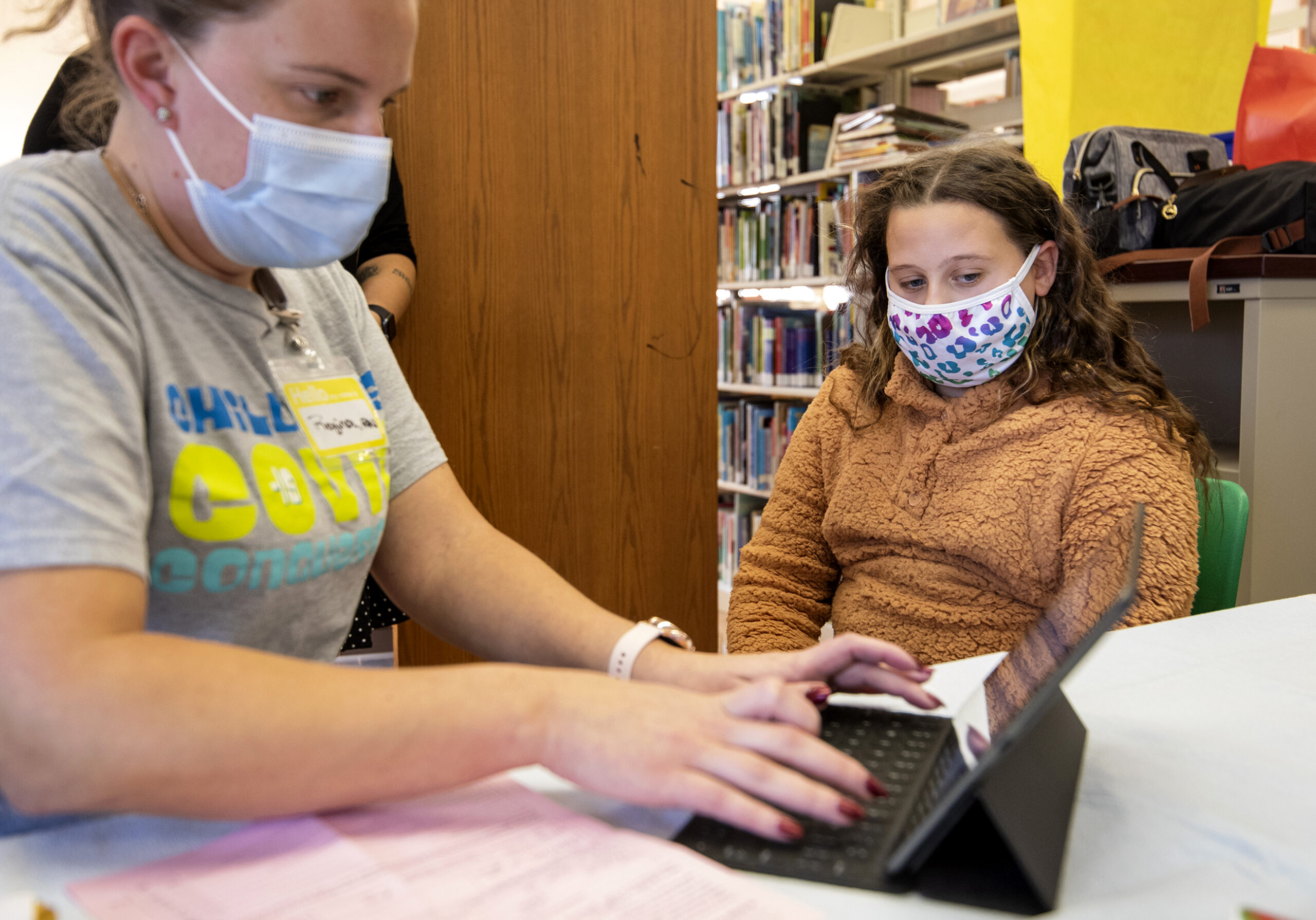 A child in a face mask watches as a nurse enters information on a laptop.