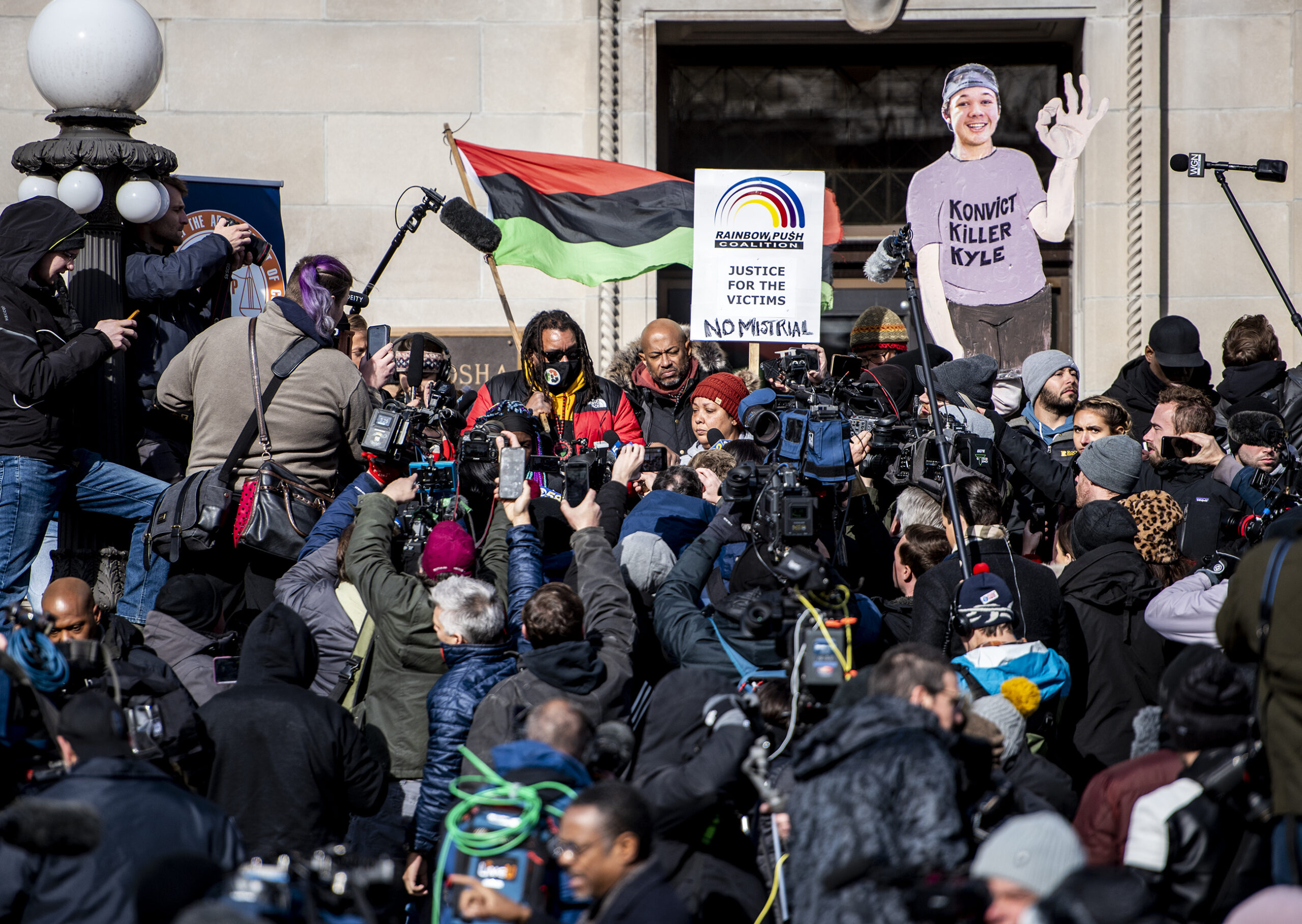 A sea of reporters and photographers take up the entire courthouse steps. Justin Blake can be seen at the top holding a flag.