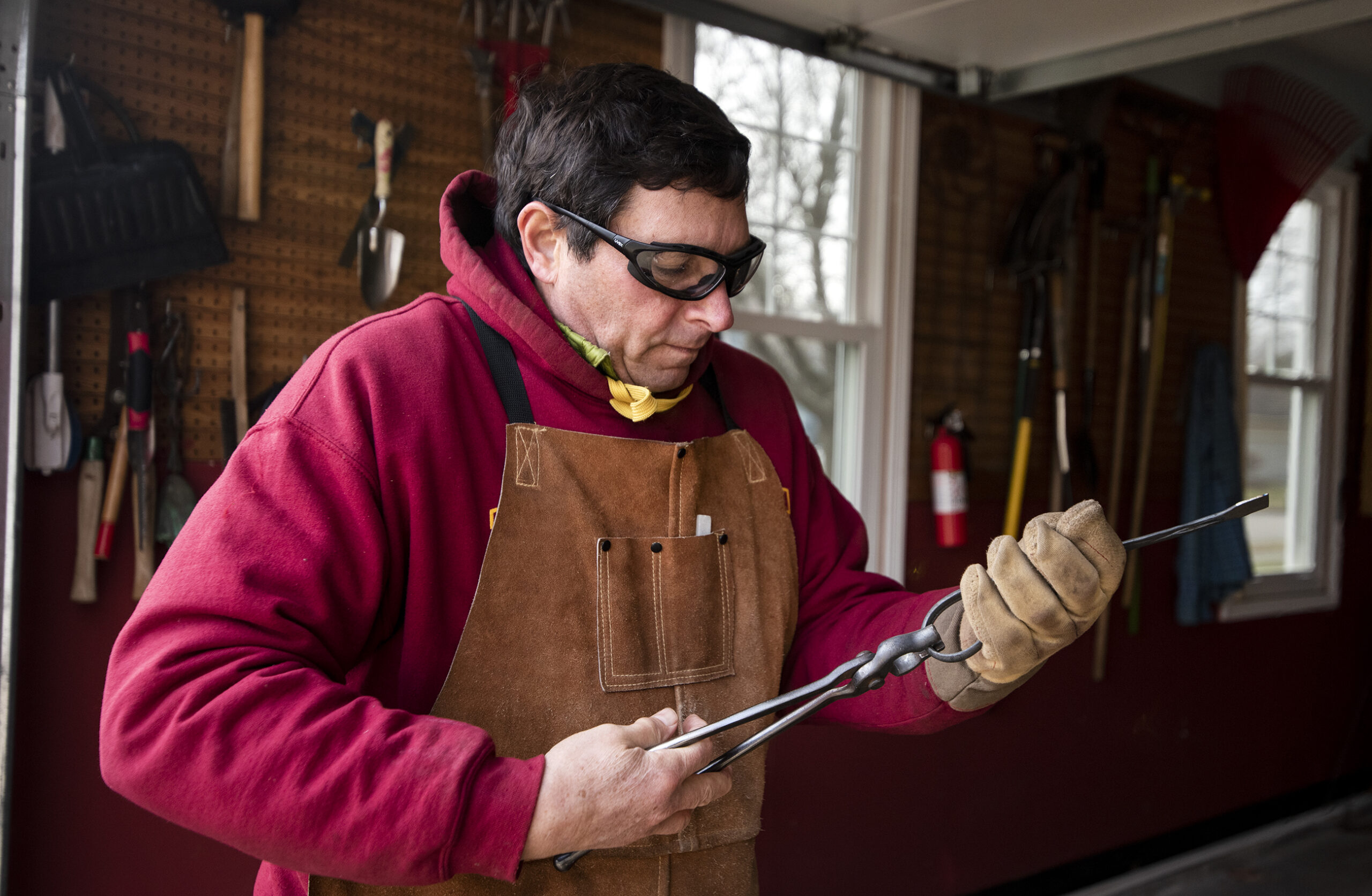 A man holds a piece of metal while wearing goggles and gloves in his garage.