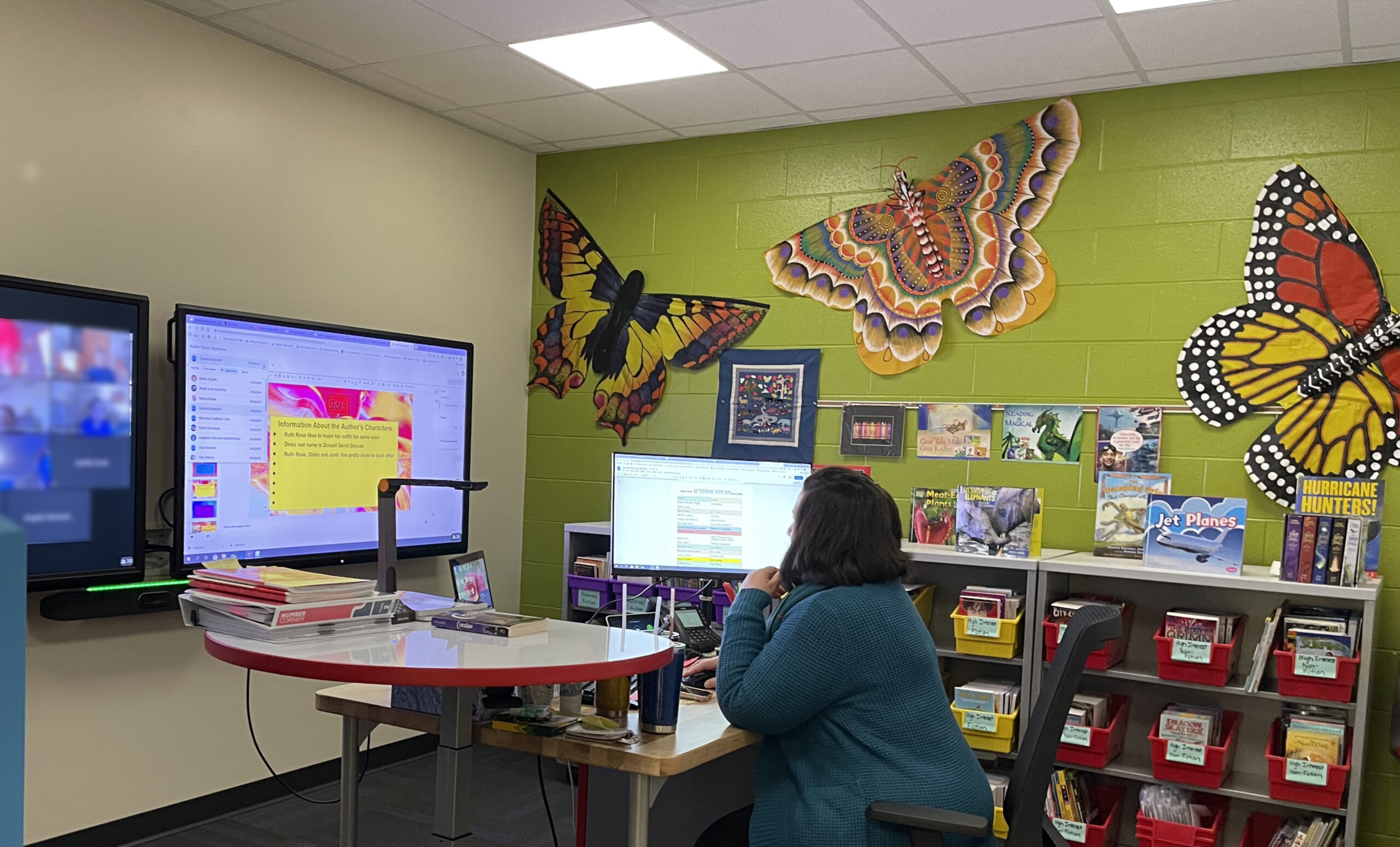 A teacher works on a computer monitor. Ahead of her, on the wall, are two TV screens, the left one with students on a video Zoom call and the right one with a school lesson.