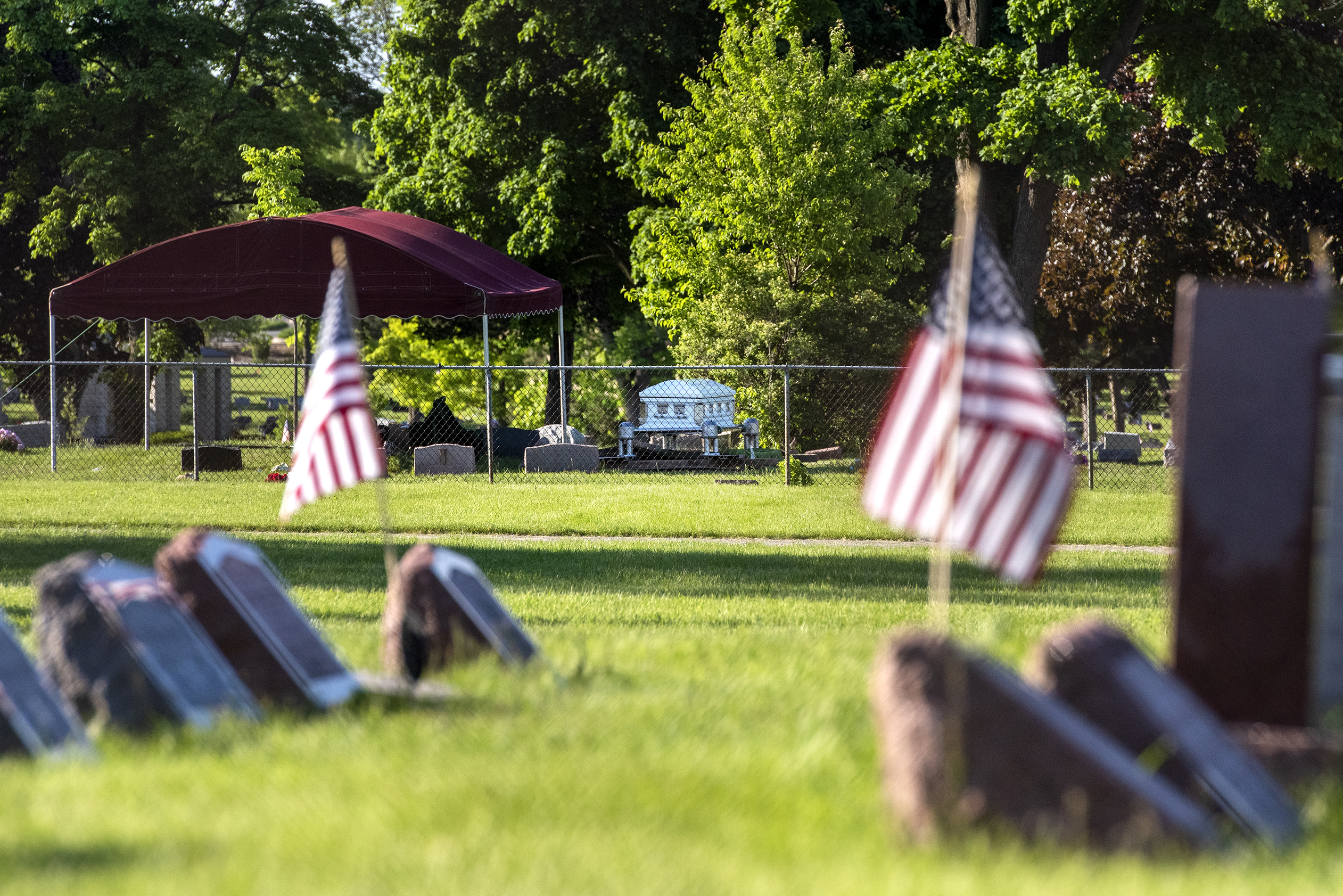 A white casket is visible outside in a cemetery. American flags adorn other gravestones nearby.