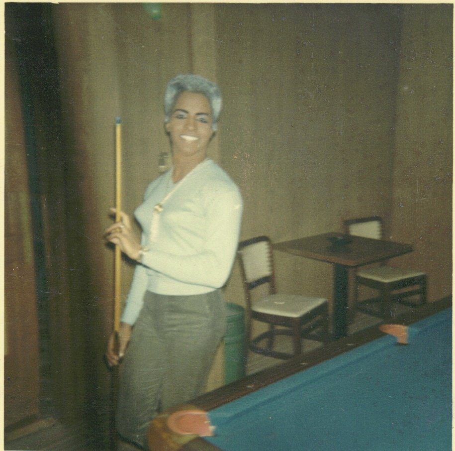 A woman of trans experience stands inside a bar next to a pool table holding a pool cue
