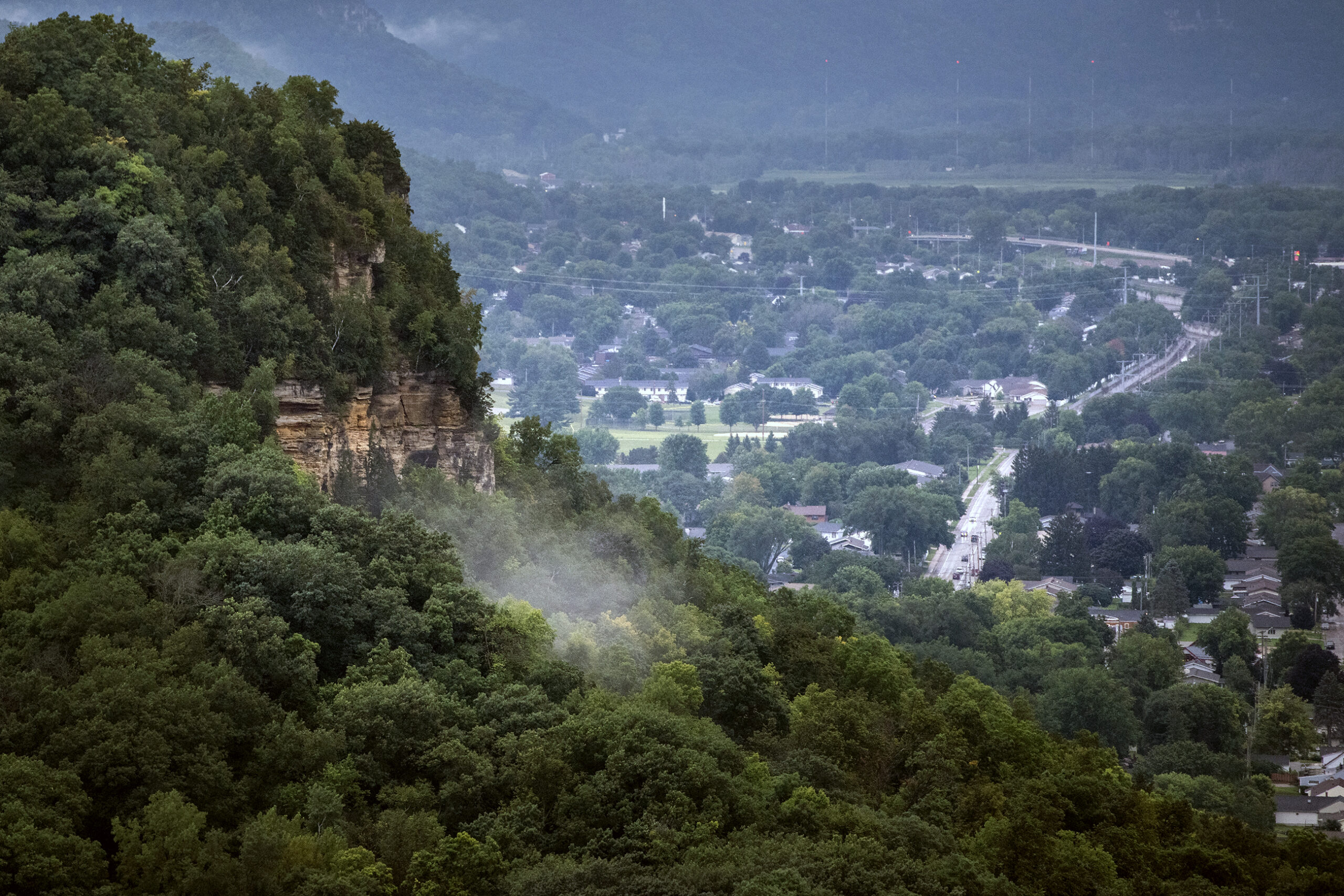 A cloud of mist on the side of a hill with green trees is seen high above the city landscape below.