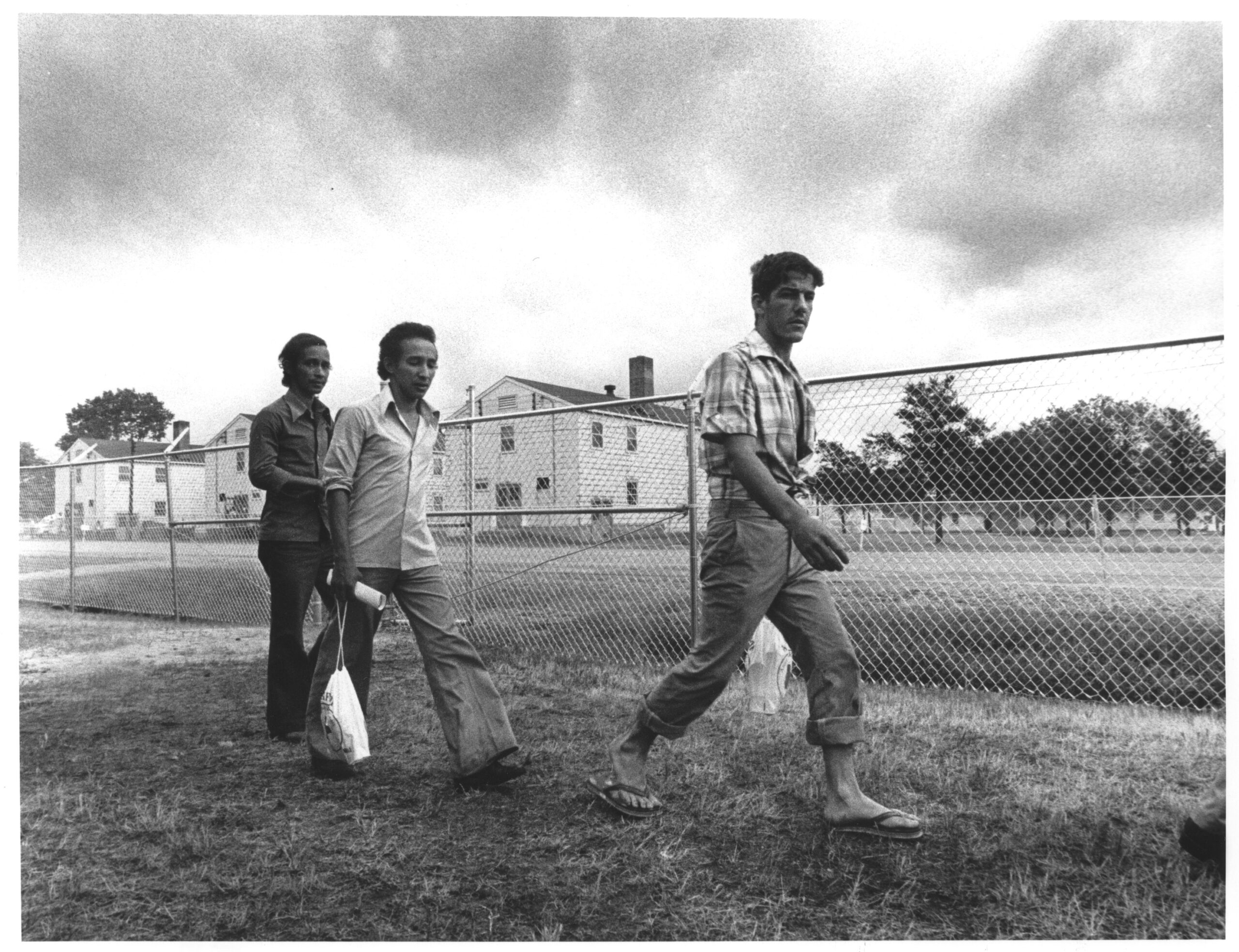 A black and white photograph featuring unidentified refugees walking along a fence