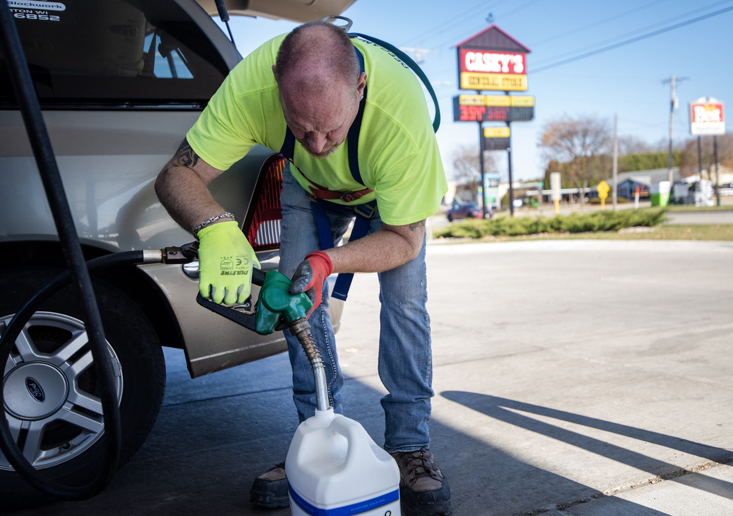 A man leans forward and fills up a tank sitting on the ground. A sign with gas prices can be seen behind him.