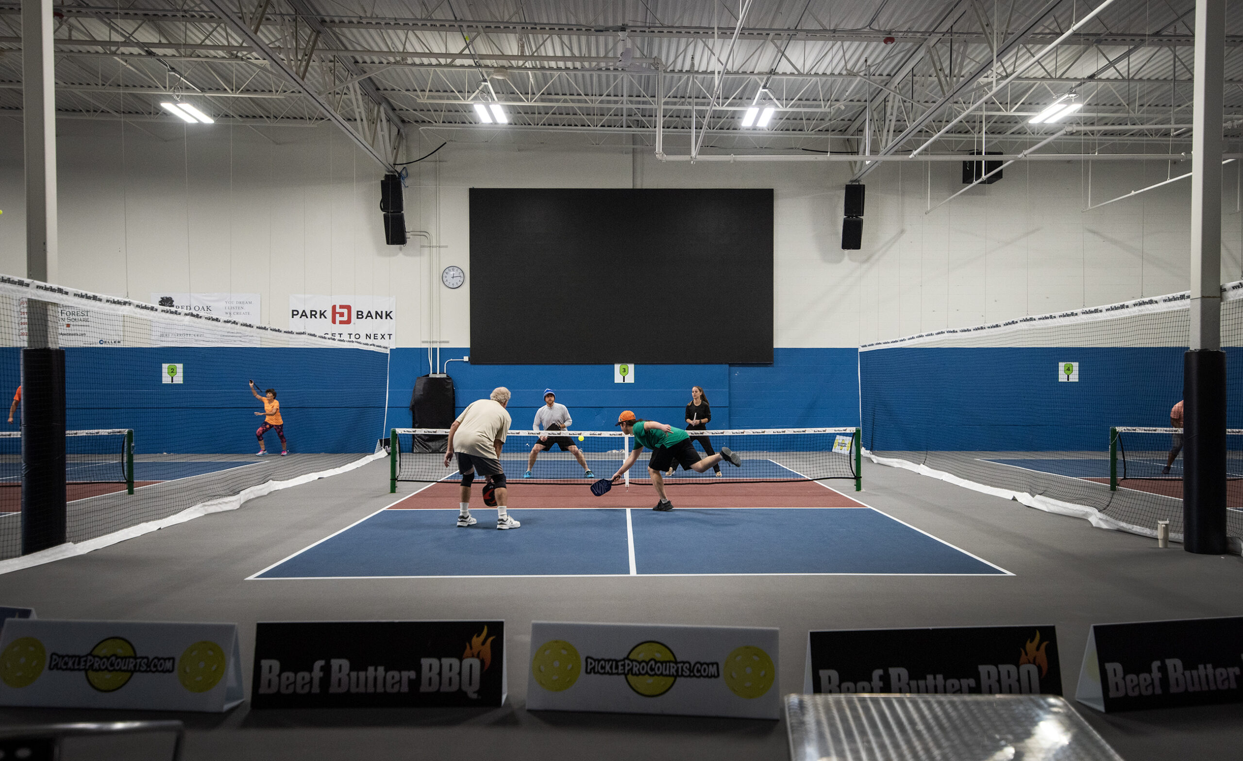 Four pickleball players play on a blue court in an indoor facility.