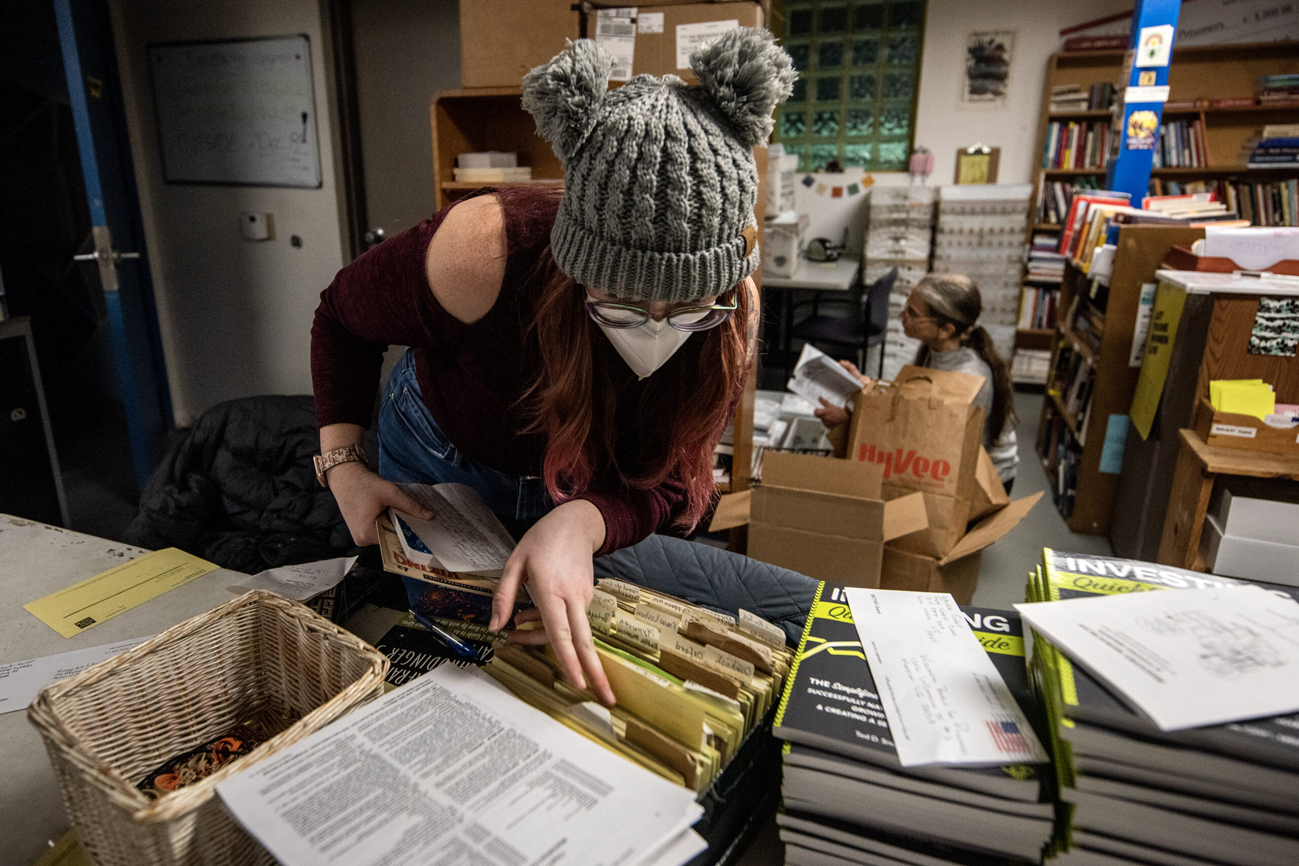 A volunteer leans over to see small papers in a box in a room stocked with books.