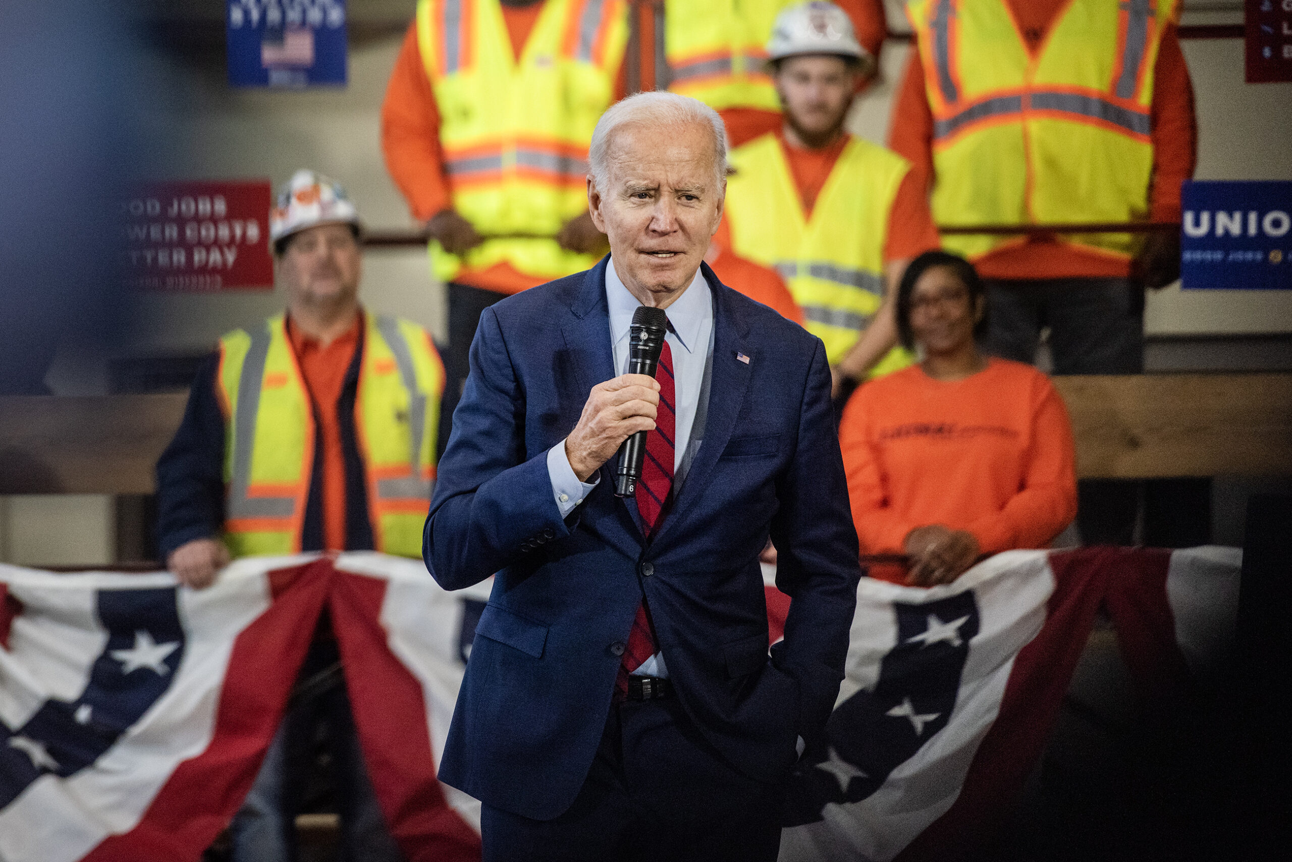 President Joe Biden speaks into a microphone. Workers in neon vests are seen behind him.