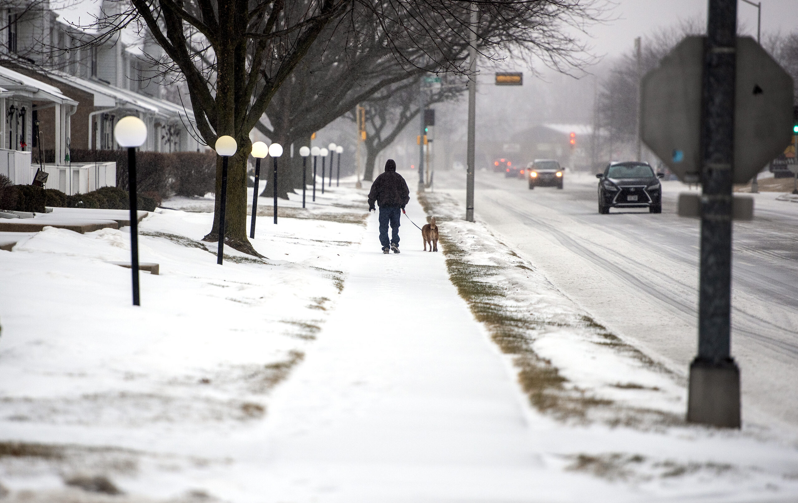 A person in a coat walks a dog on a leash across a snowy sidewalk.