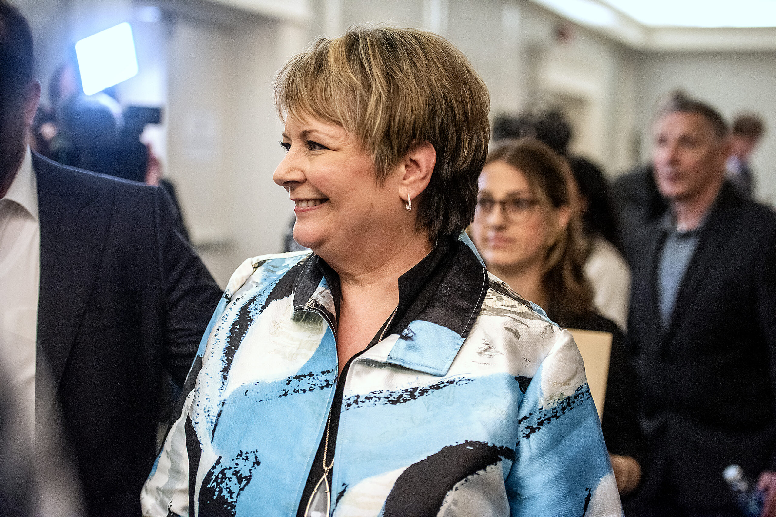 Janet Protasiewicz wears blue and white and smiles as she walks through the room.