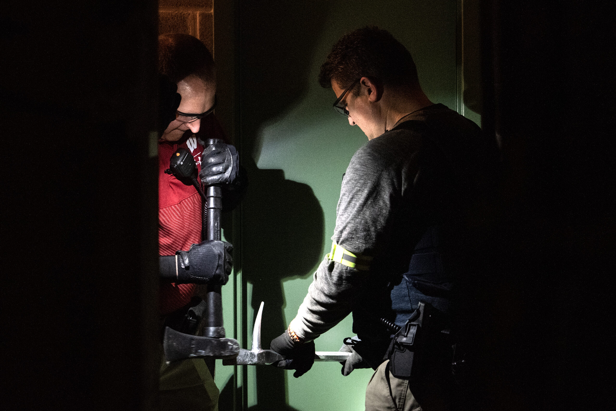 Two officers work in the dark to pry open a door with large metal tools.