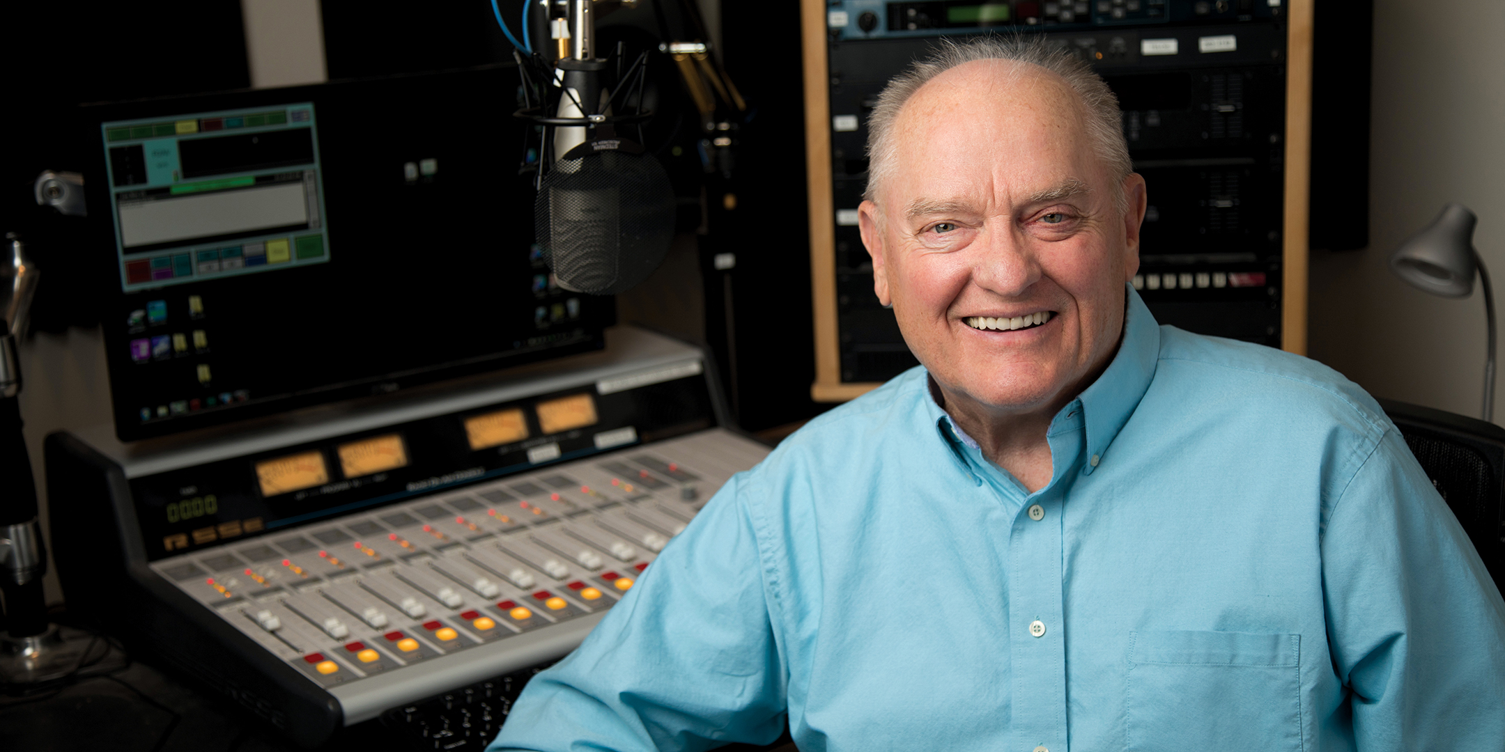 Larry Meiller sitting at a broadcast control board. He smiles warmly and is wearing a blue shirt.