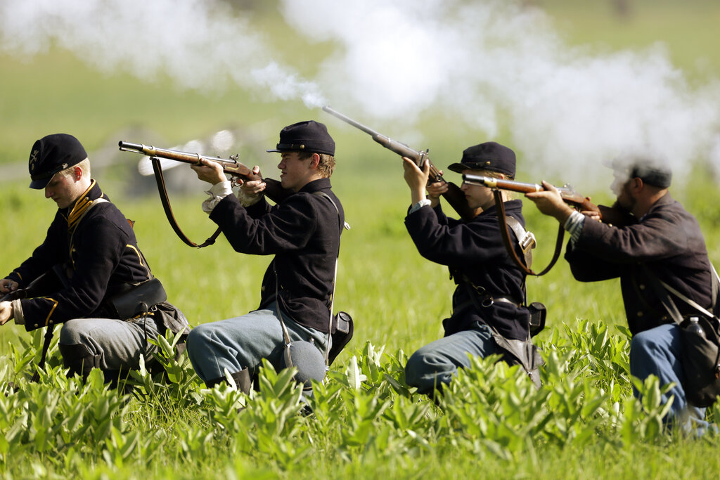 A line of people dressed in Union Civil War uniforms kneel in the grass and fire off old guns.