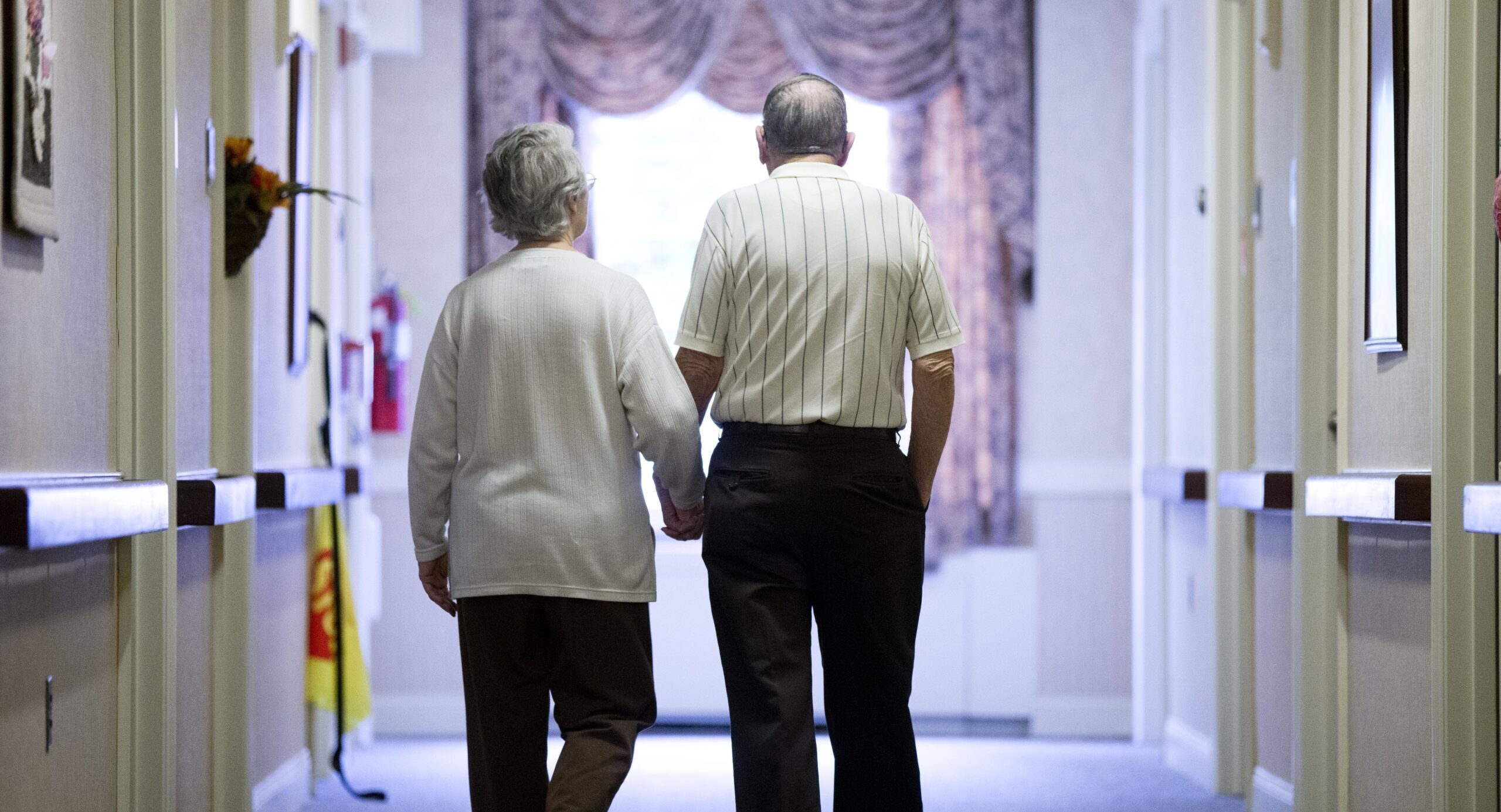 An elderly couple walks down a hall of a nursing home