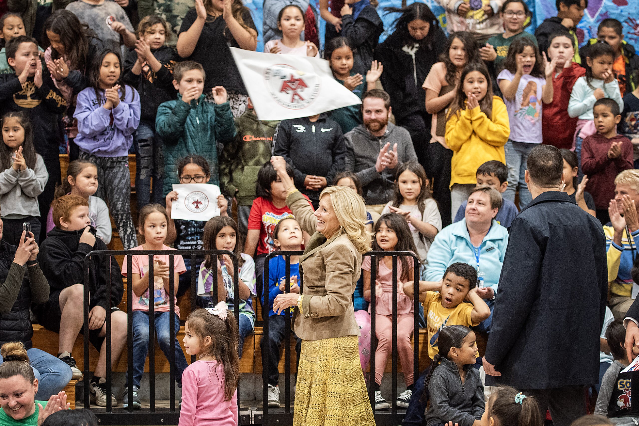 Students watch and cheer as Jill Biden waves a white flag with a seal on it.