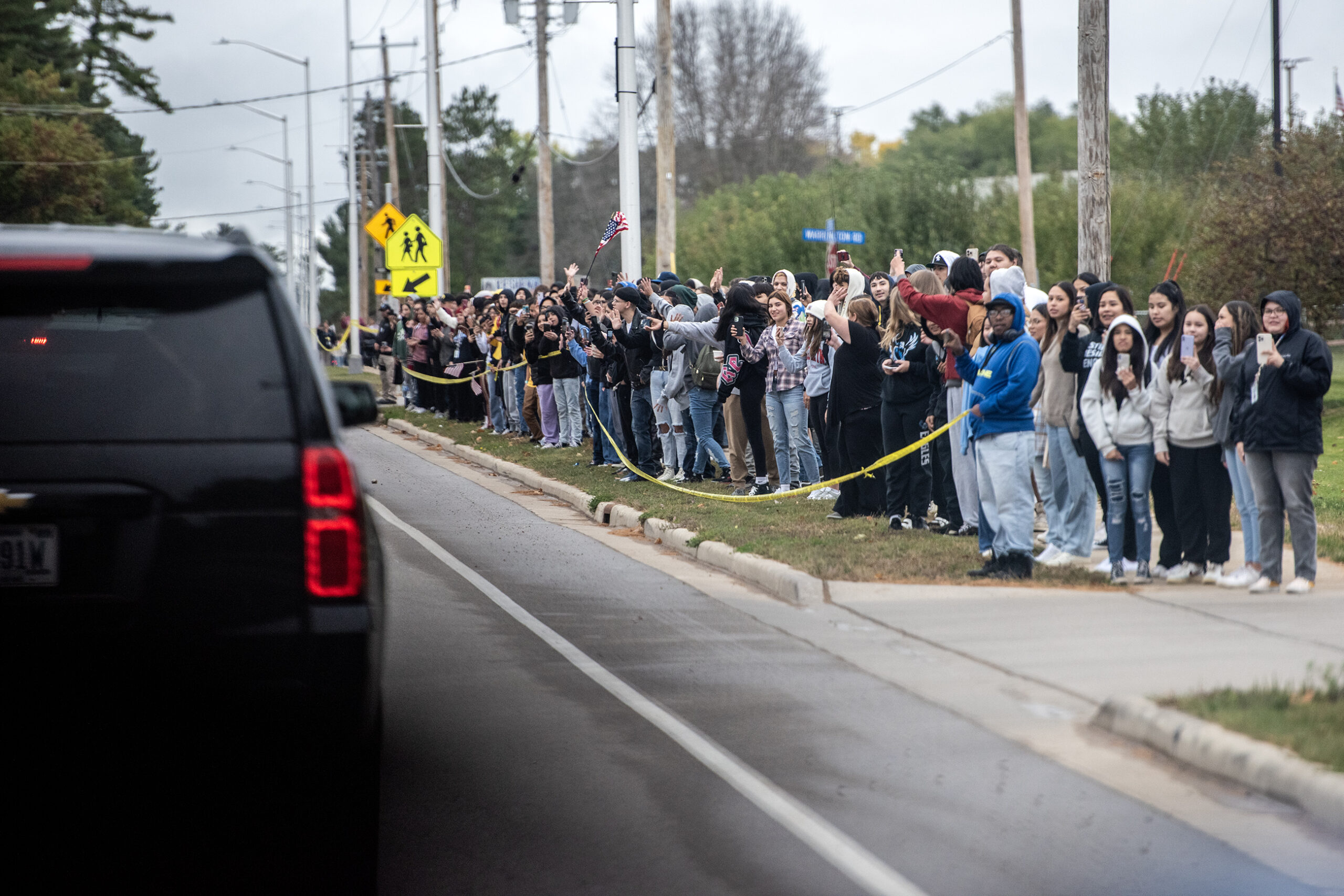 A crowd of students stand outside as black SUV's pass by.