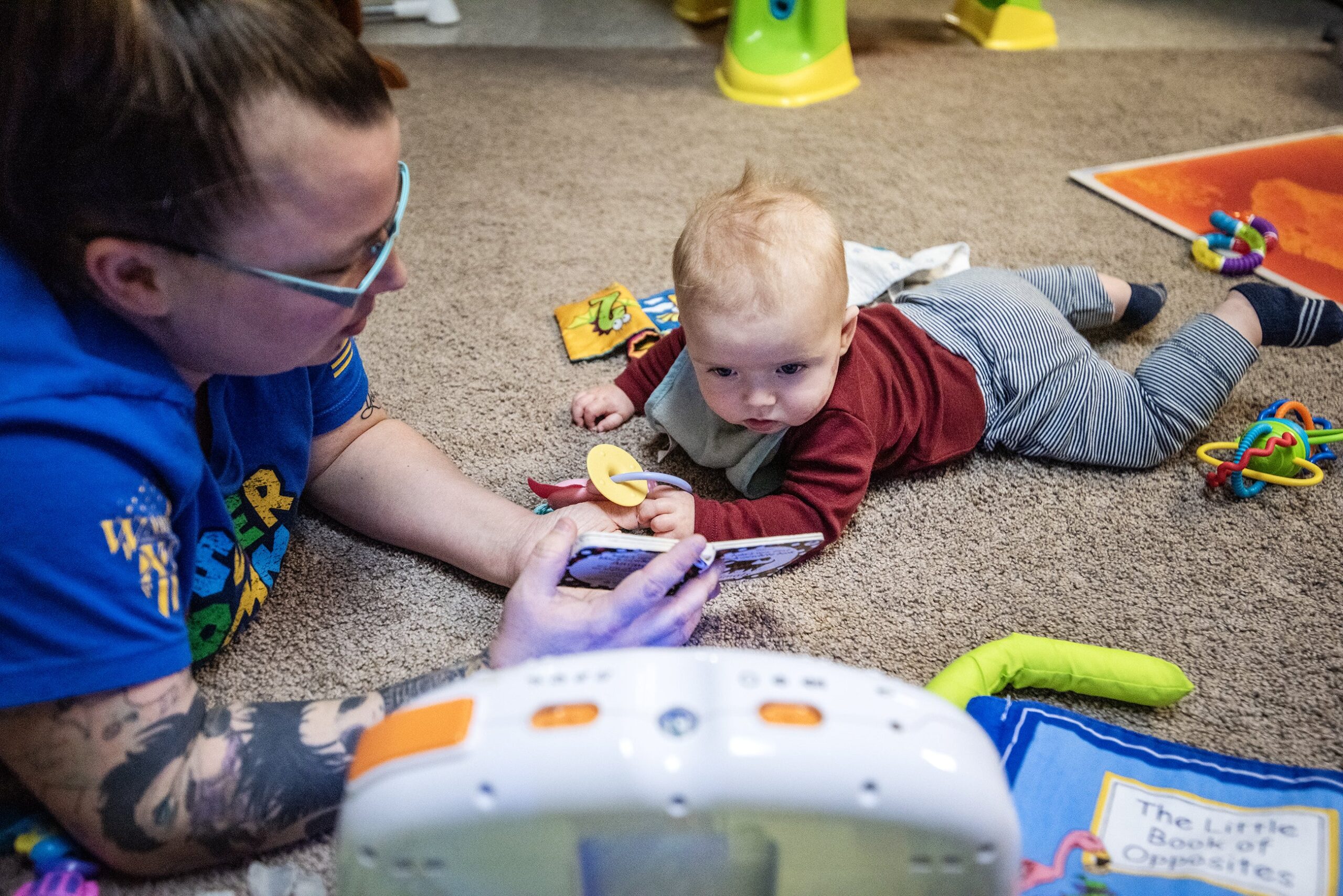 A baby lays in a play area as a teacher holds up a book.