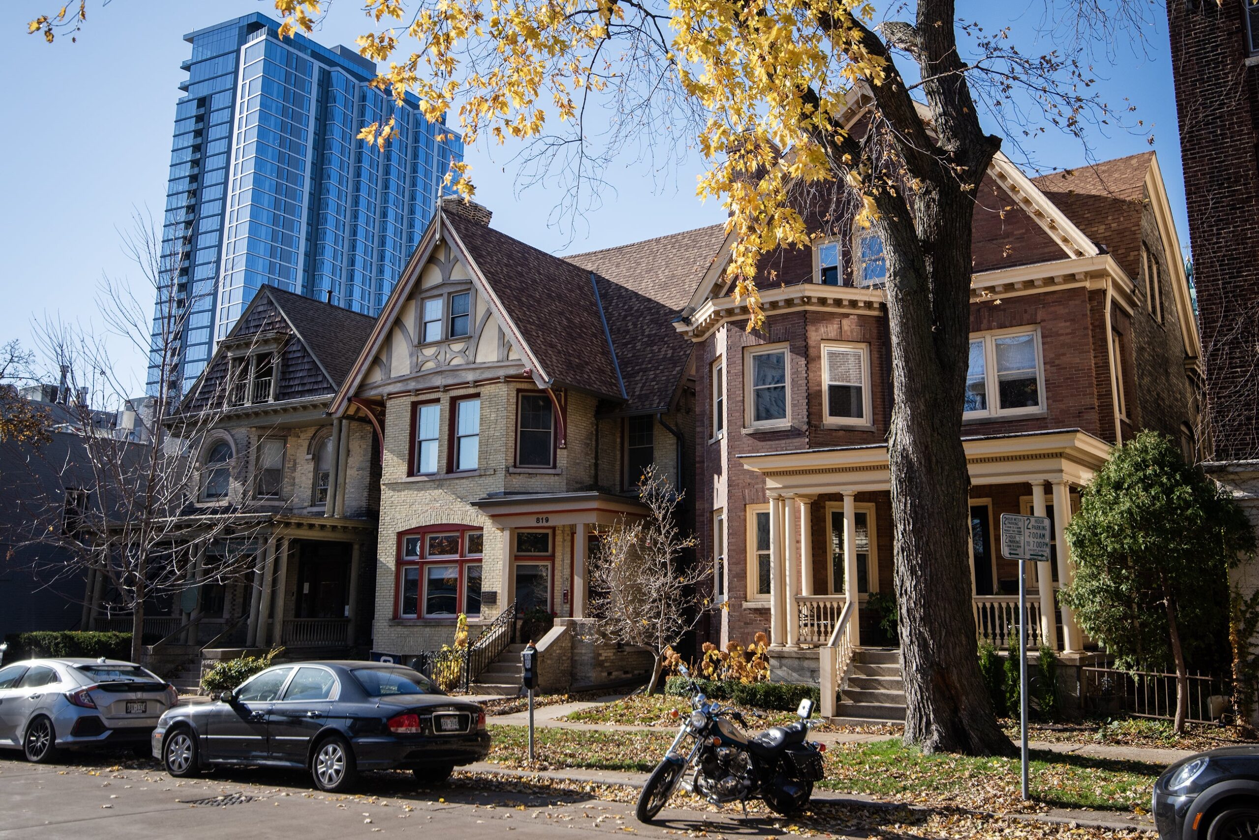 Three homes turned into rental units are side-by-side on a sunny fall day. A tall business building is in the background.