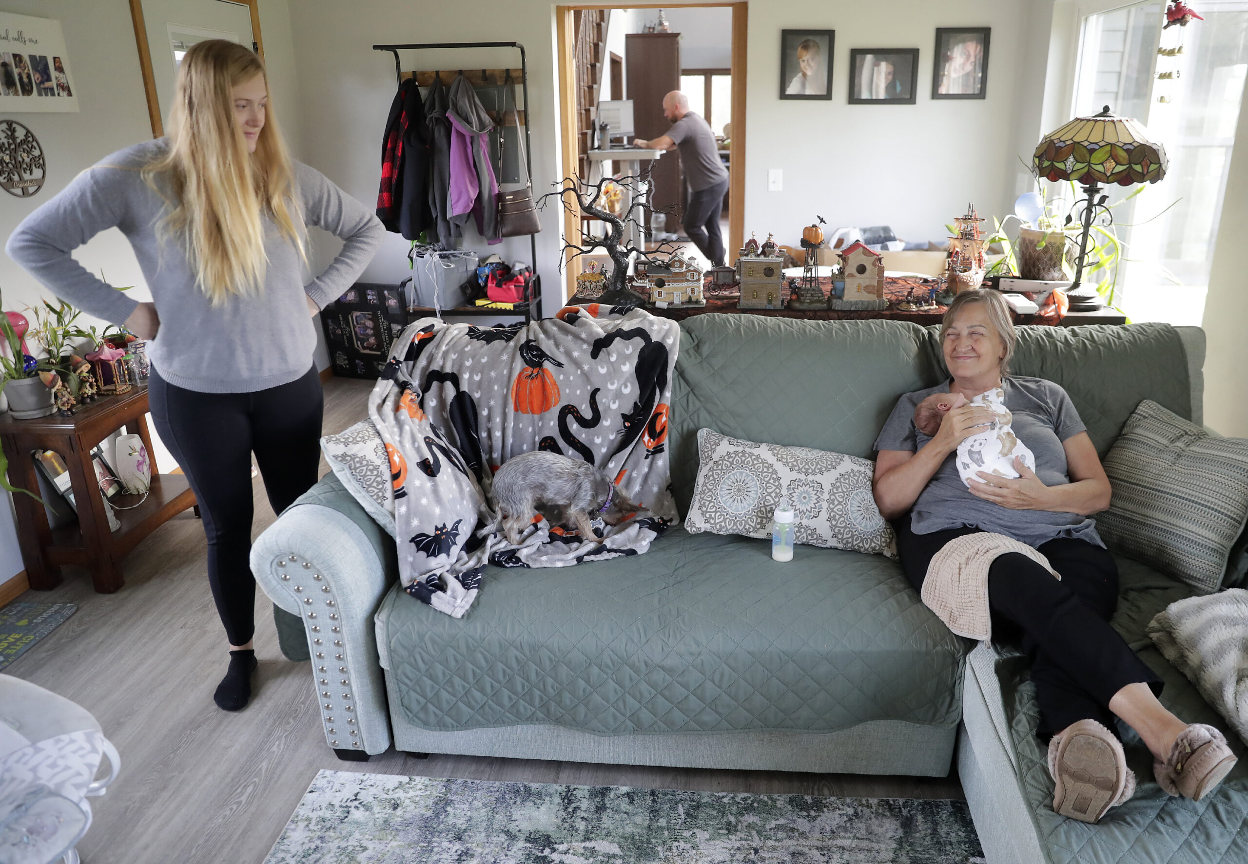 A woman sits on the couch with her grandson while his mother stands in the living room next to them