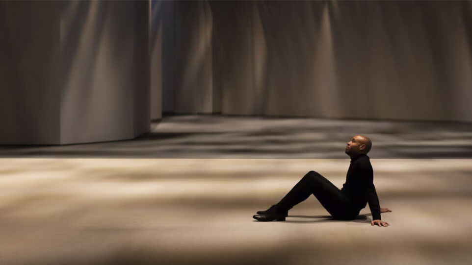 Aaron Diehl is clad all in black. He sits on the floor of a bare stage, leaning back on his hands. He's looking up.