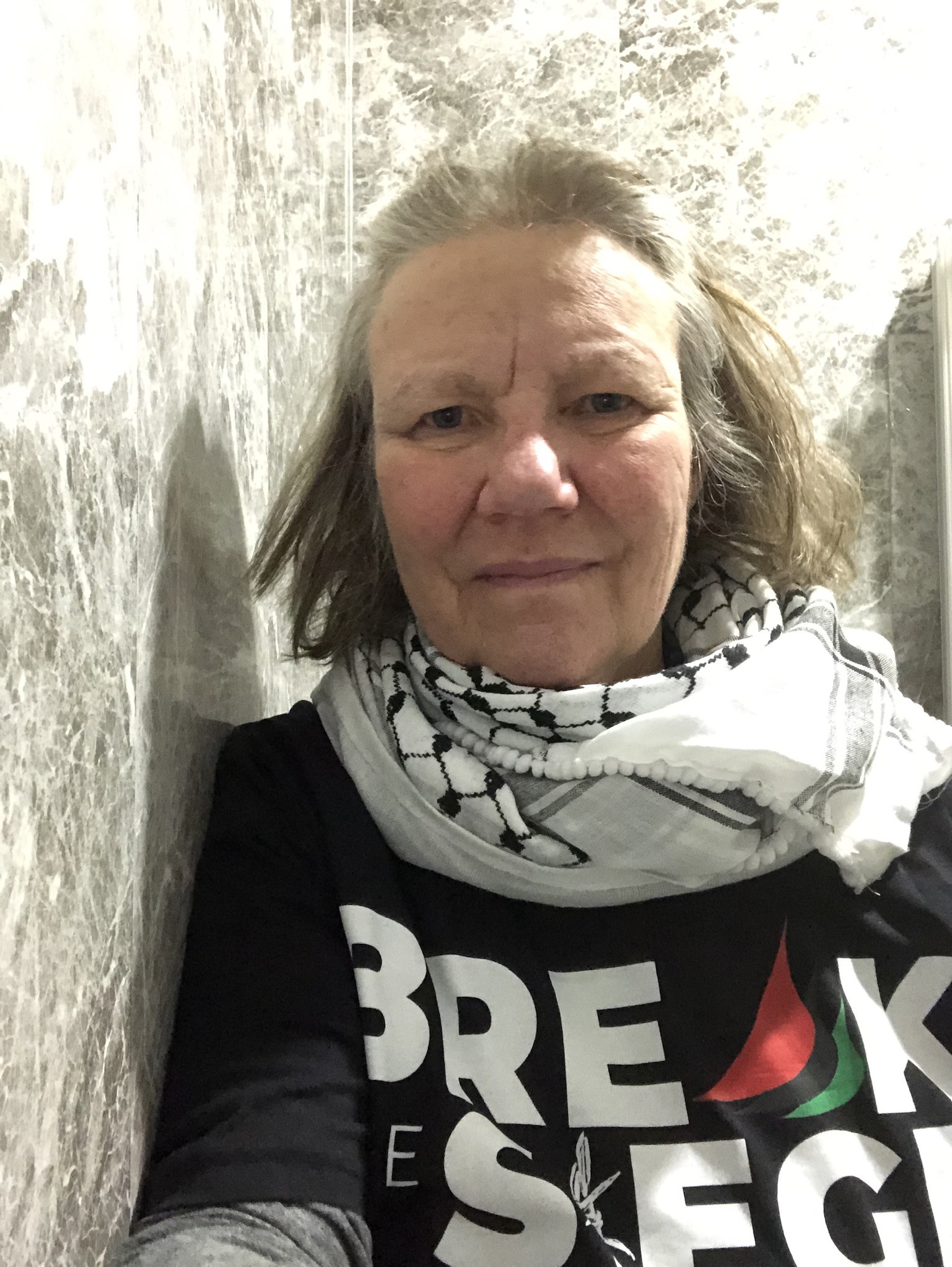 A woman looks directly at the camera while wearing a white and black scarf and a shirt reading "Break the Siege"