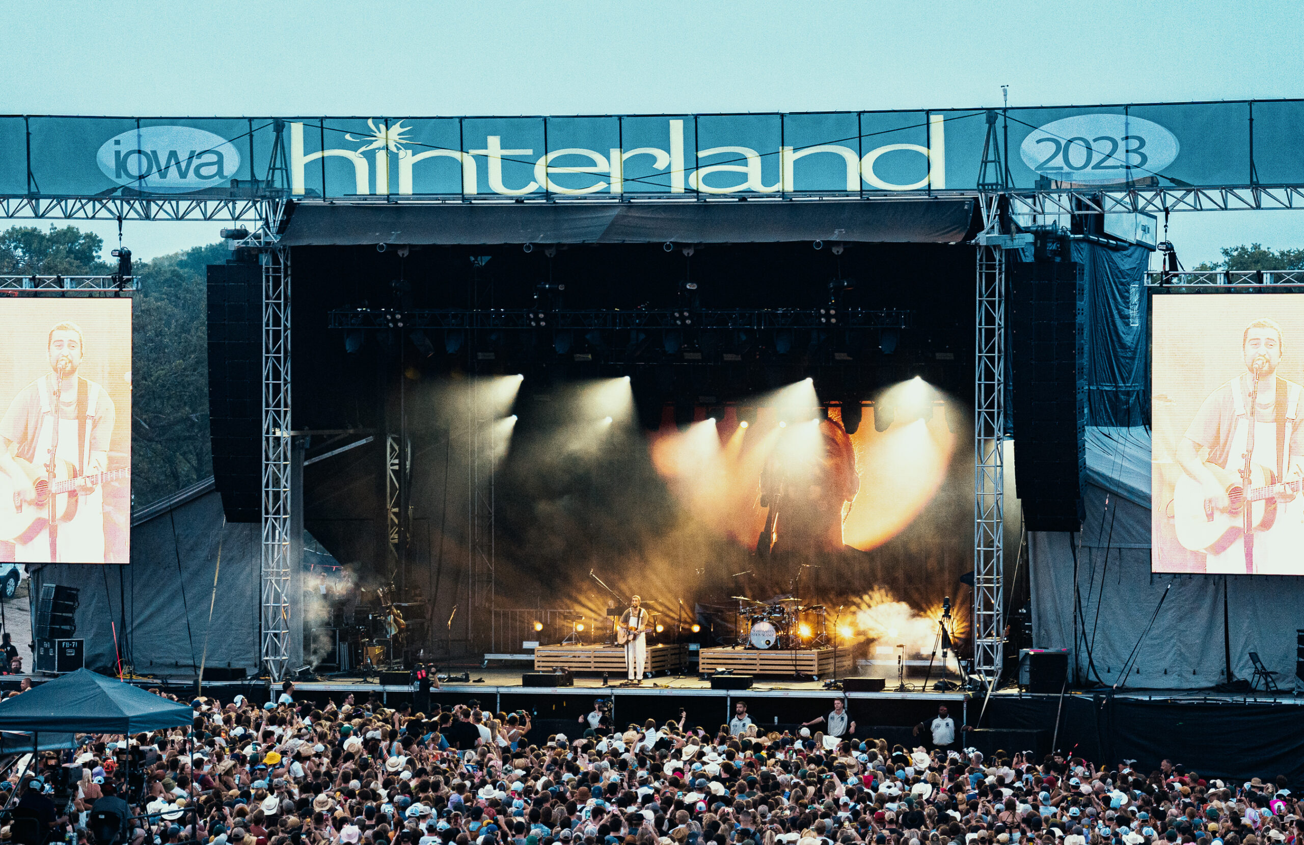 Noah Kahan performs on the Hinterland stage in front of a large crowd in 2023. A Hinterland sign is above the stage in blue with with letters.