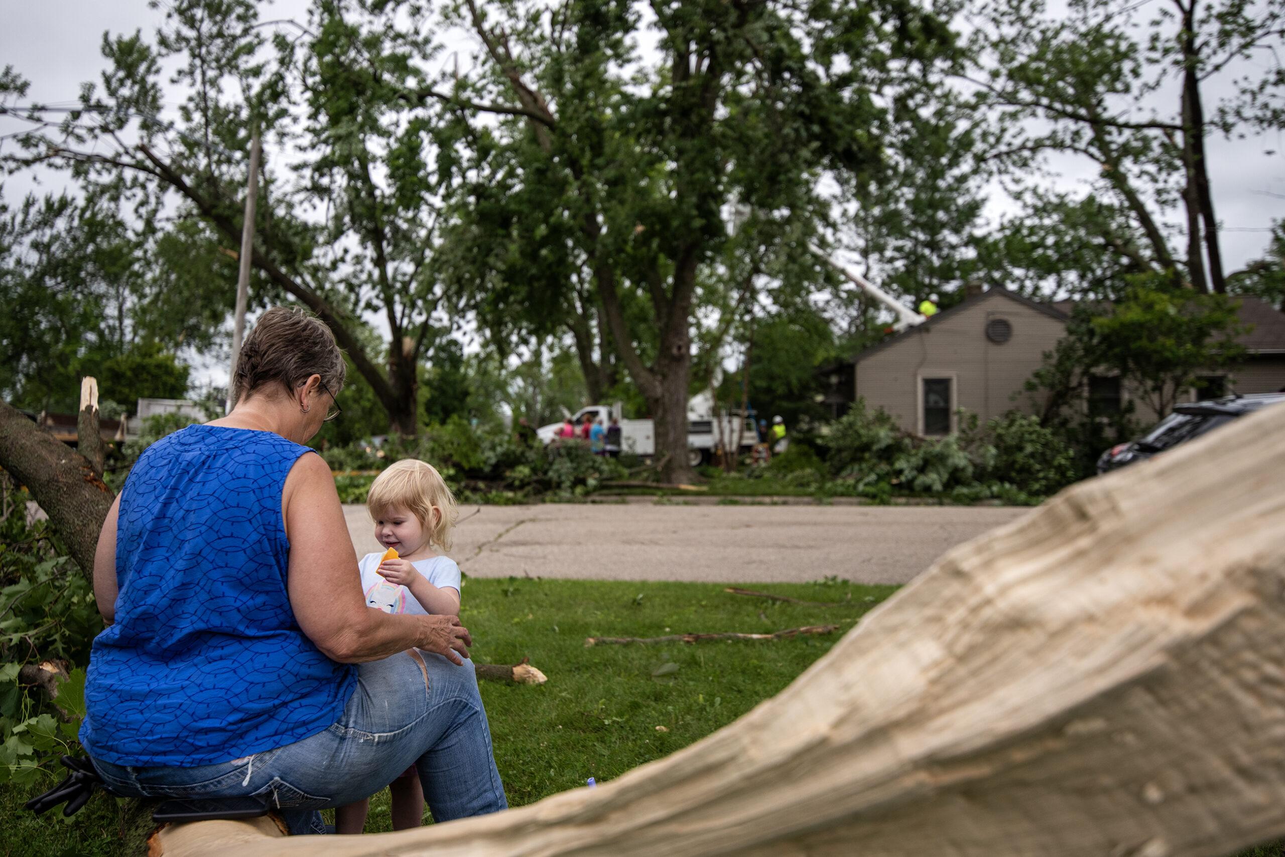 A woman and a chid sit together on a broken tree branch near storm damage.