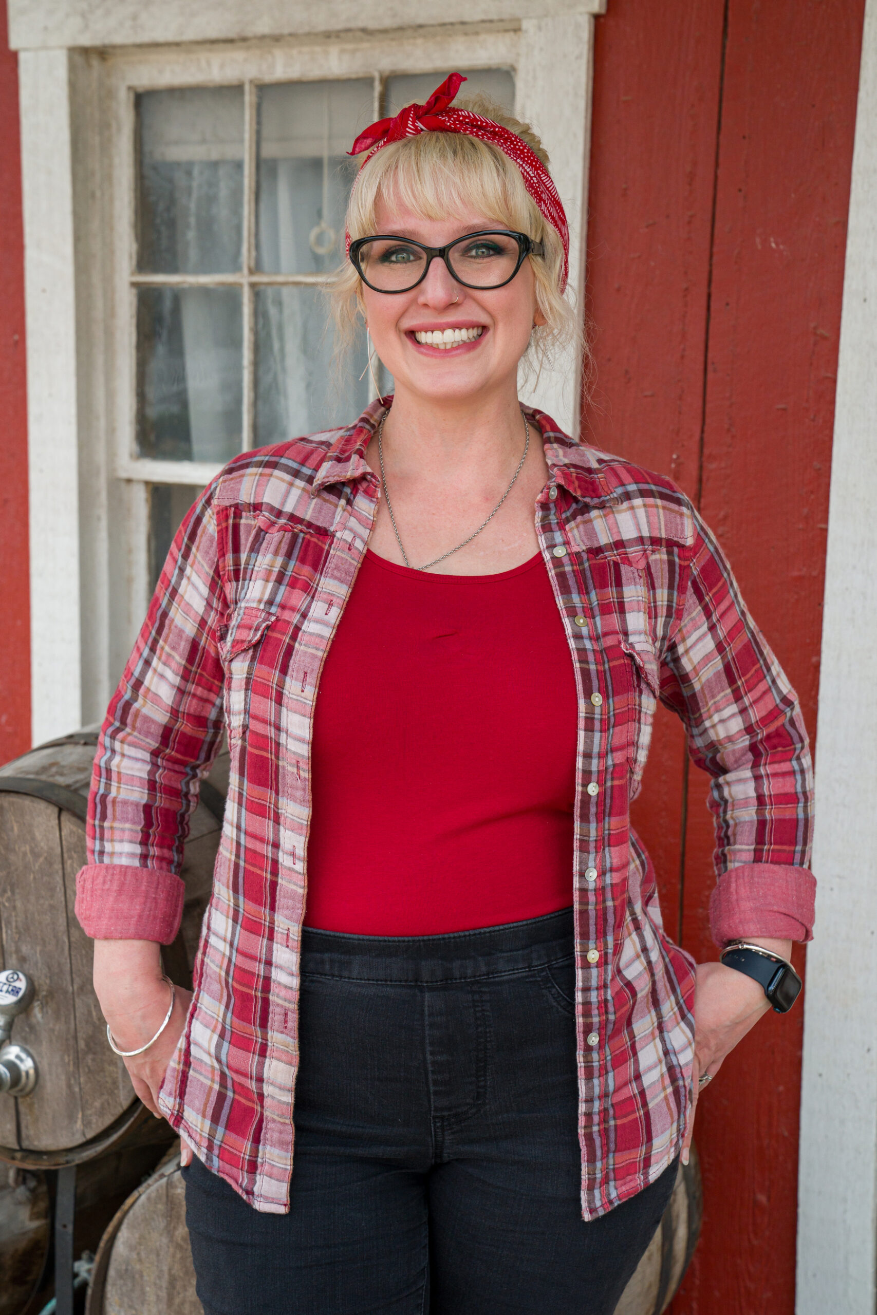 A woman with blonde hair, glasses, and a plaid shirt smiles in front of a rustic red building