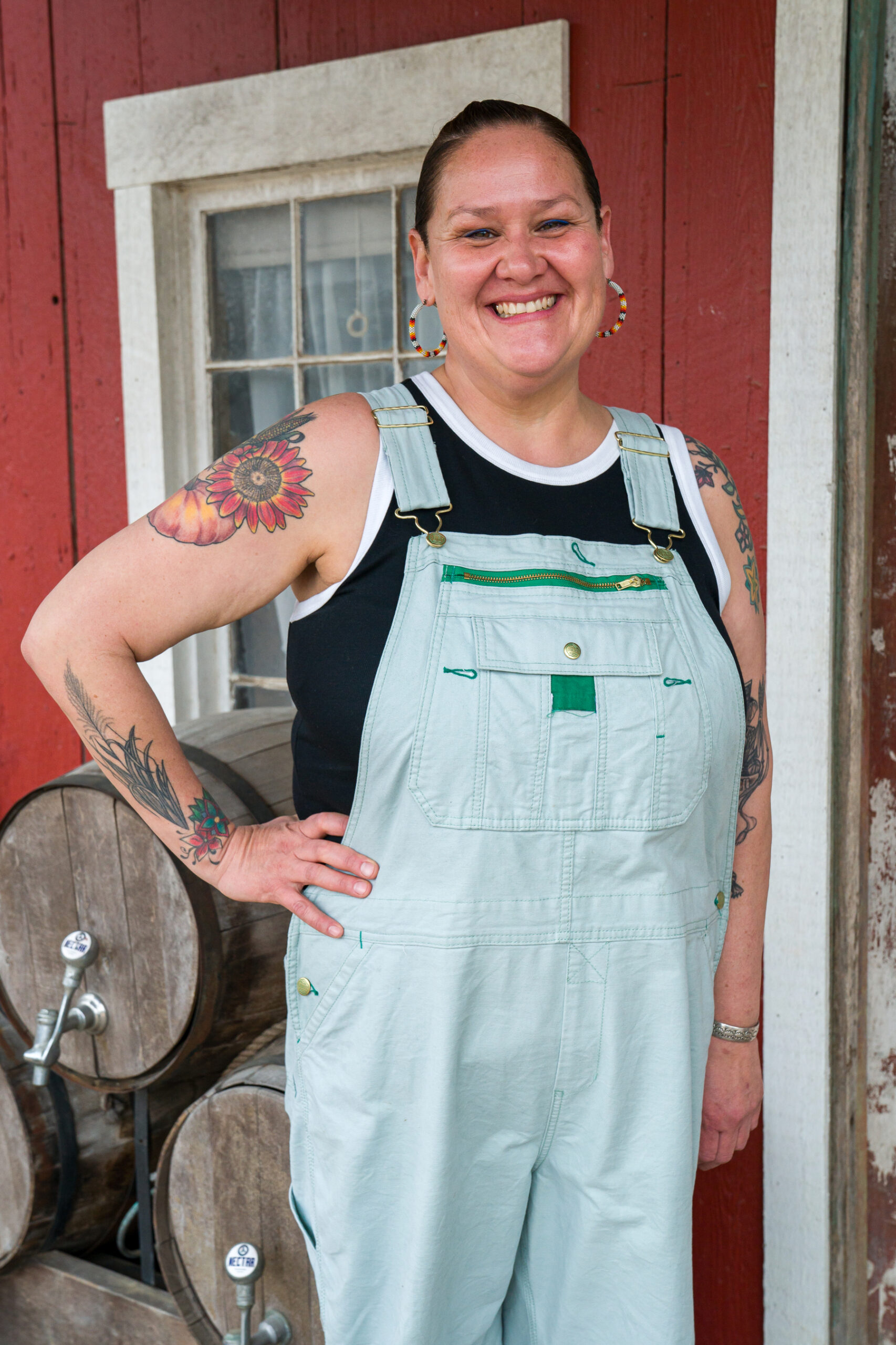 A woman wearing bead earrings and overalls smiles in front of a rustic red building