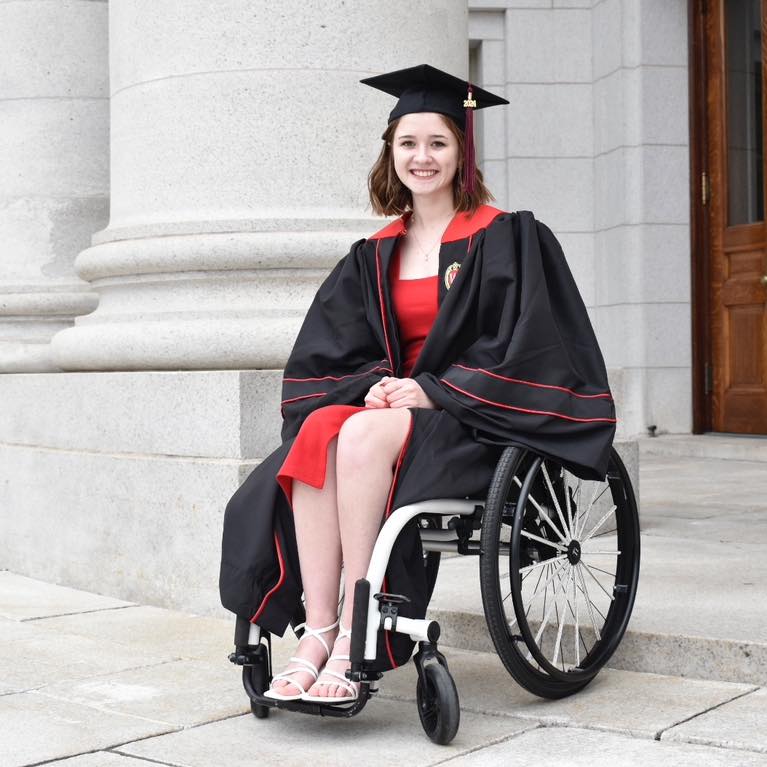 A young woman poses in a wheelchair wearing a black and red cap and gown in front of a large stone building