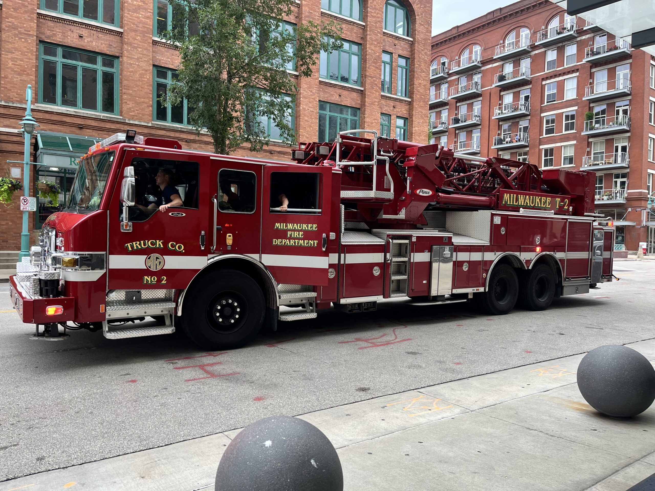 A red firetruck from the Milwaukee Fire Department is parked on a road in front of two brick apartment buildings.