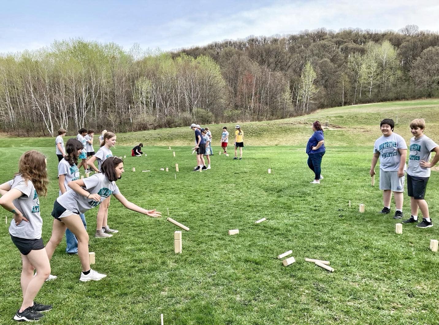 Kids wearing gym t-shirts throw wooden kubb batons in a green field