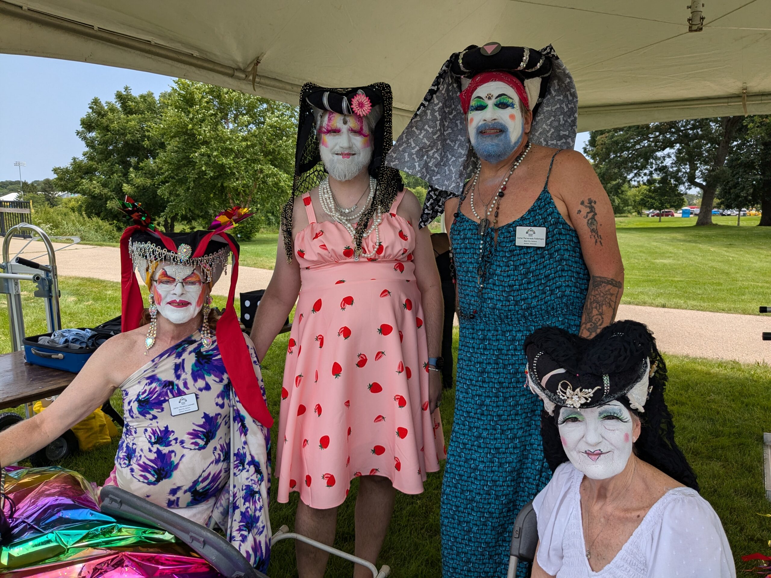 Four people in colorful makeup and dresses pose for the camera under an event tent in a park