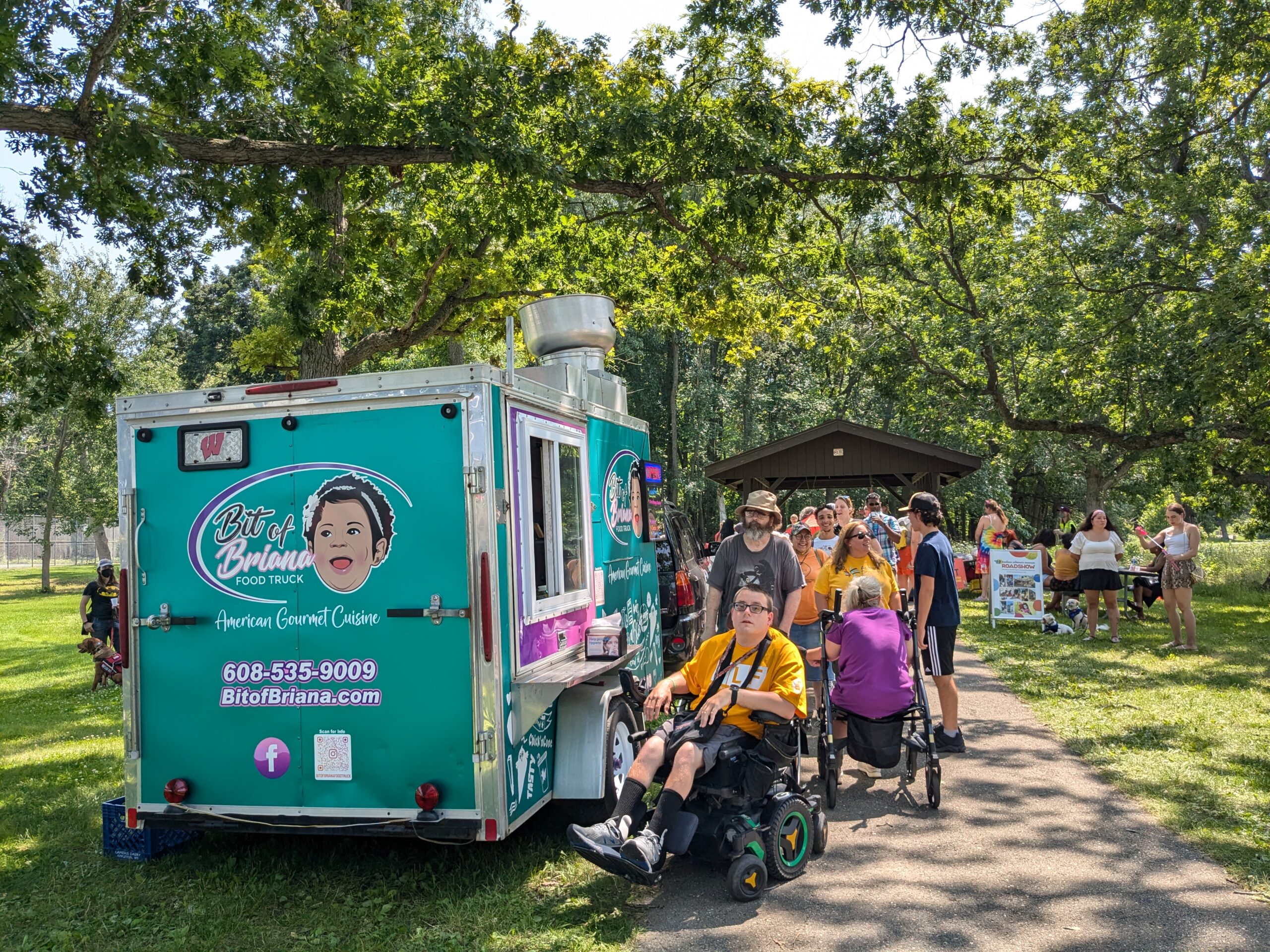 A crowd of people stands and sits in wheelchairs in front of a colorful blue-green food truck with the logo "Bit of Briana."