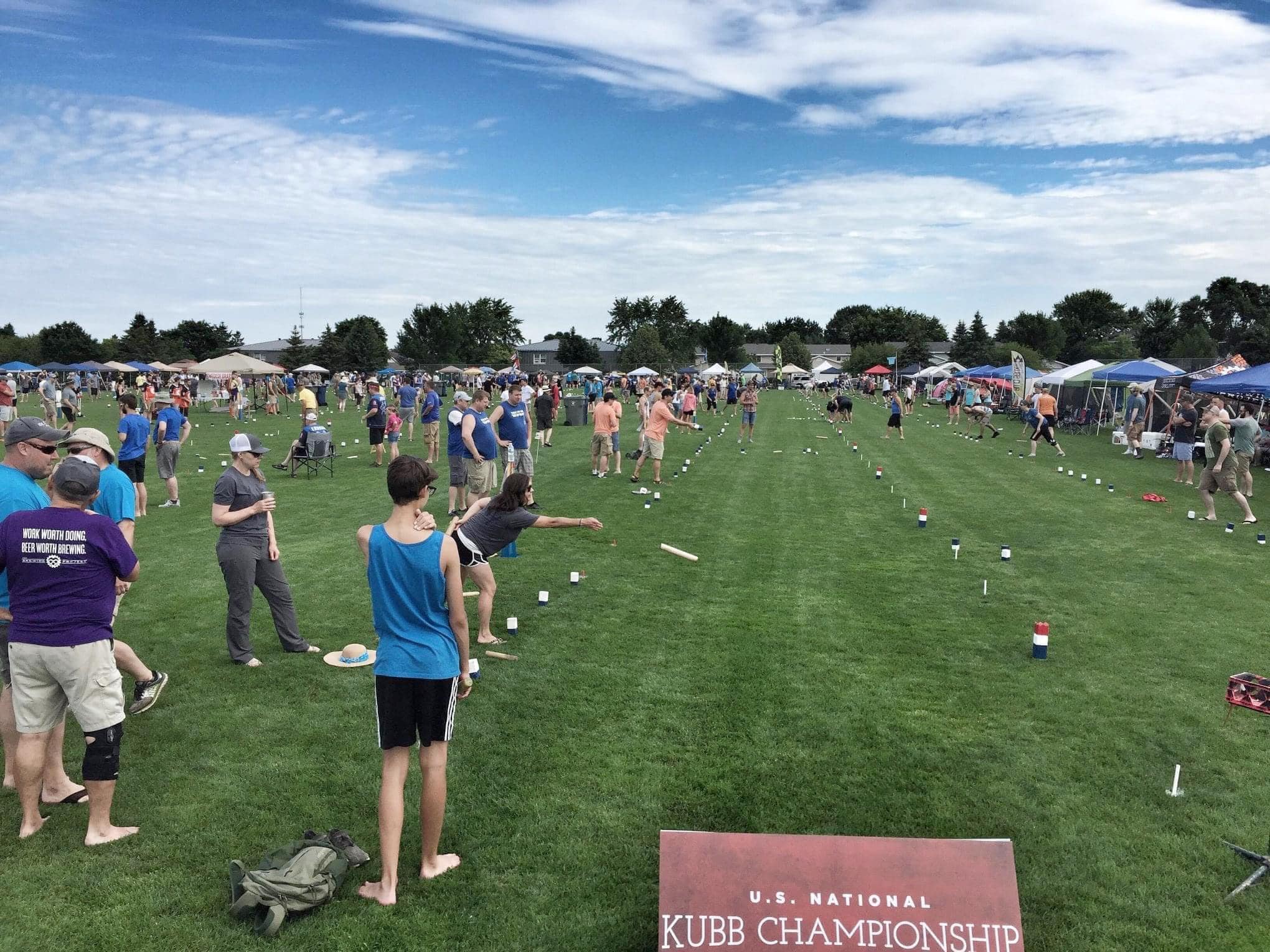 A large group of people is standing on a soccer field with event tents and a sign reading "U.S. National Kubb Championship"