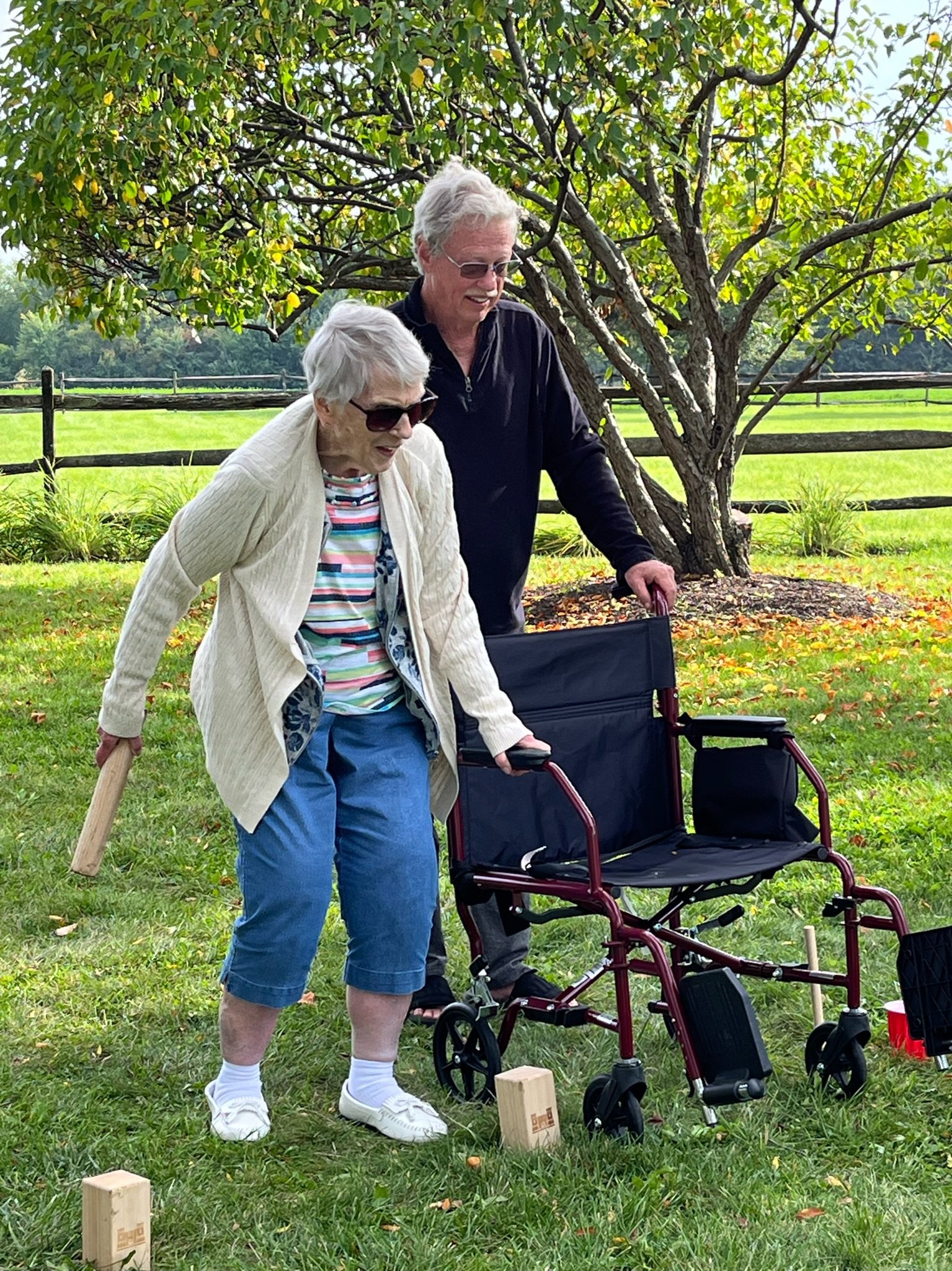An elderly woman holding onto a wheelchair bends her knees while preparing to toss a wooden kubb baton