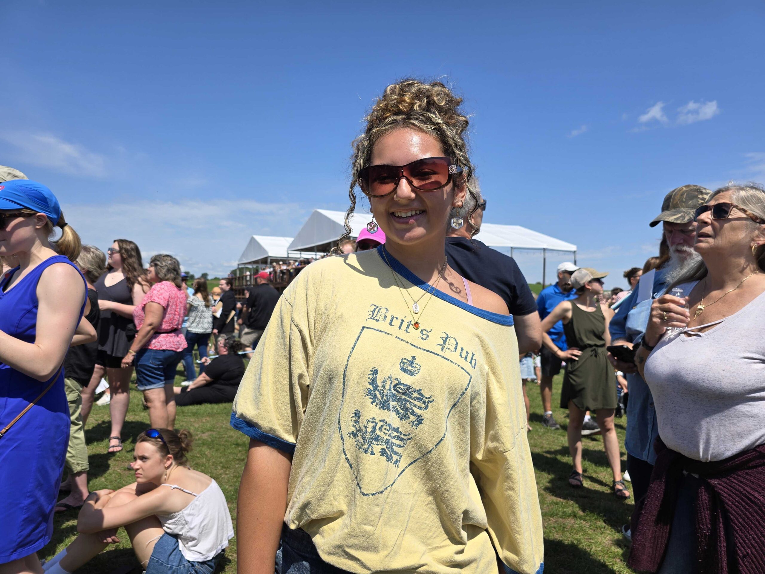 A 17-year-old girl in a yellow Brit's Pub t-shirt smiles at a Kamala Harris rally in Eau Claire, Wis. She is in sunglasses with a blue sky behind her, as well as other attendees.