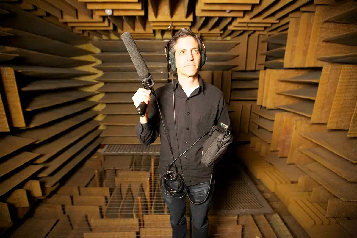 A man stands in a room surrounded by soundboards, holding a shotgun microphone