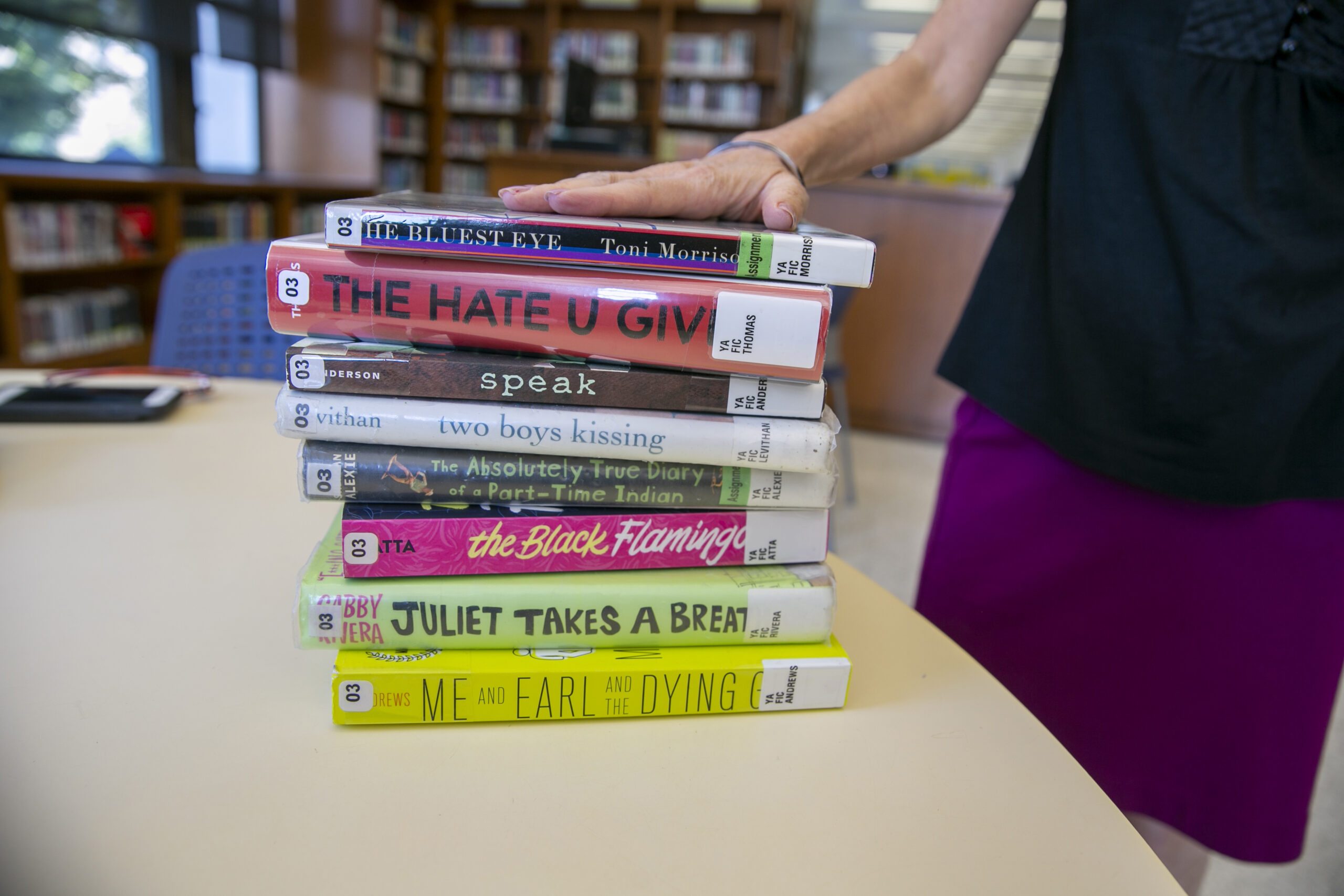 A hand rests atop a pile of books with colorful jackets and library catalog tags