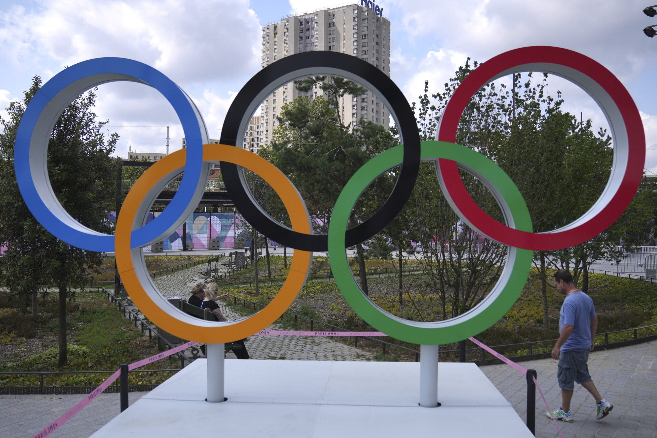 The olympic rings in front of a tall concrete building and some trees.