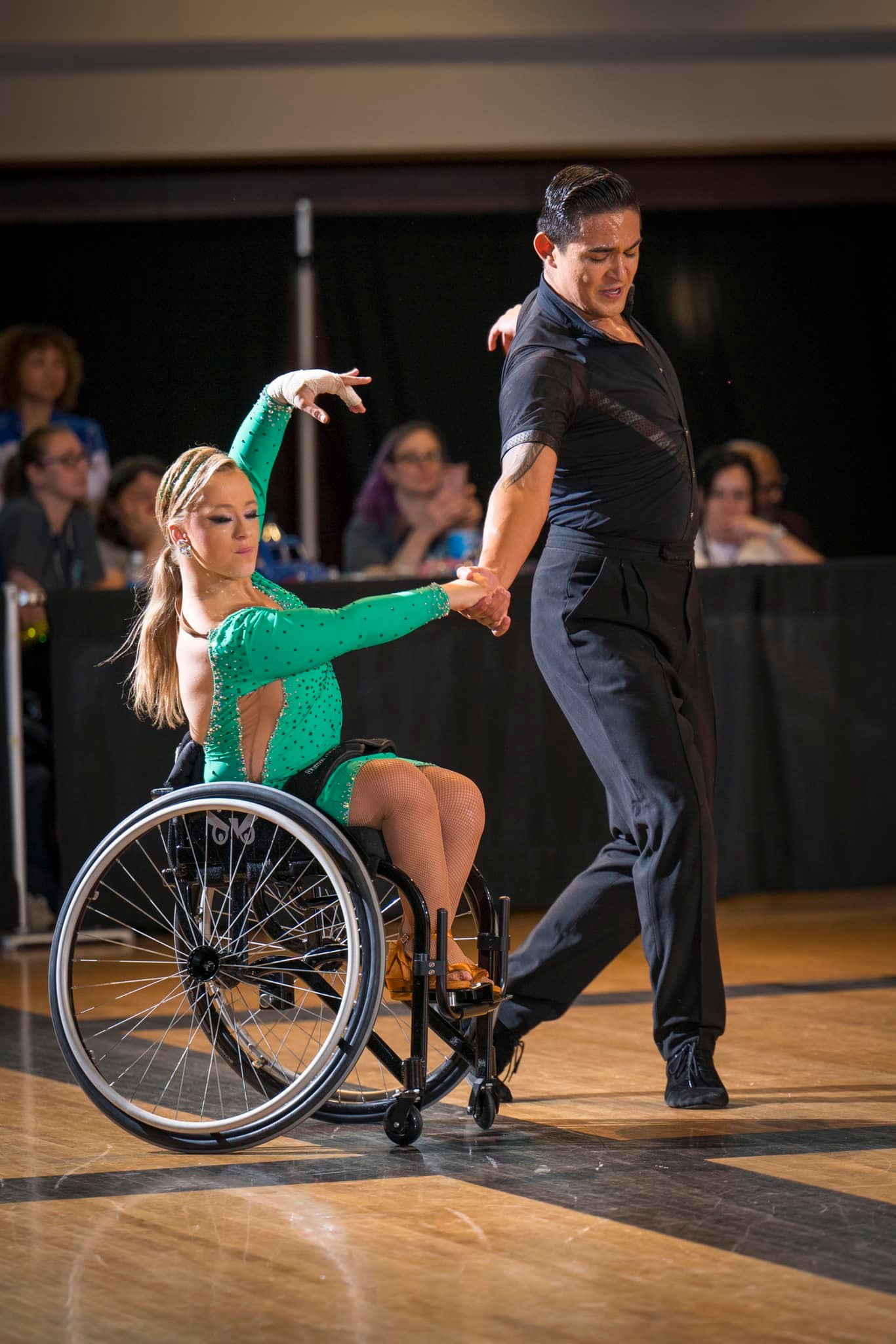 A woman in a wheelchair dances in a green sequin dress with a standing partner in a black suit