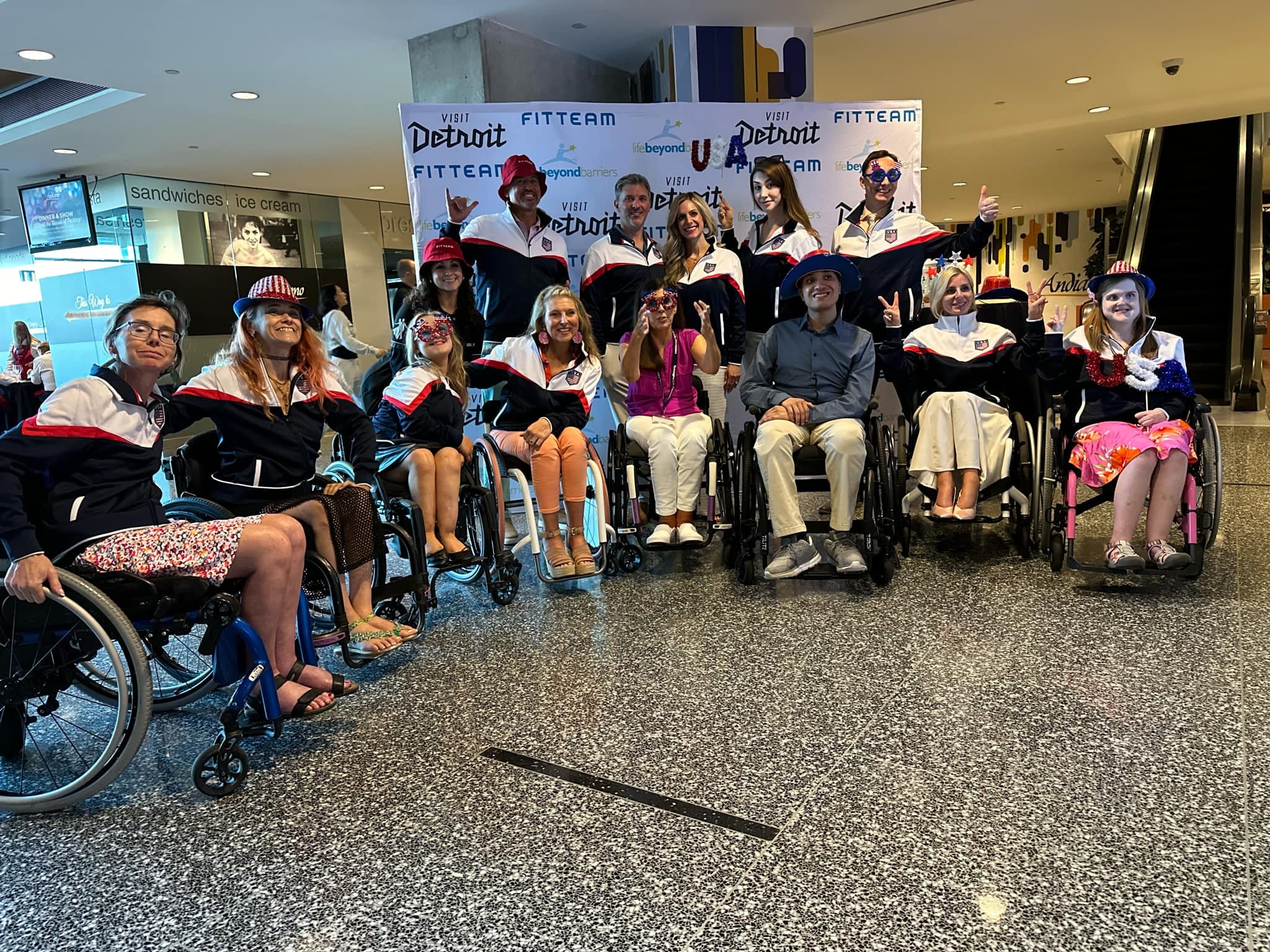 A group of eight people in wheelchairs and six standing people pose together in jackets, hats and glasses with USA motifs